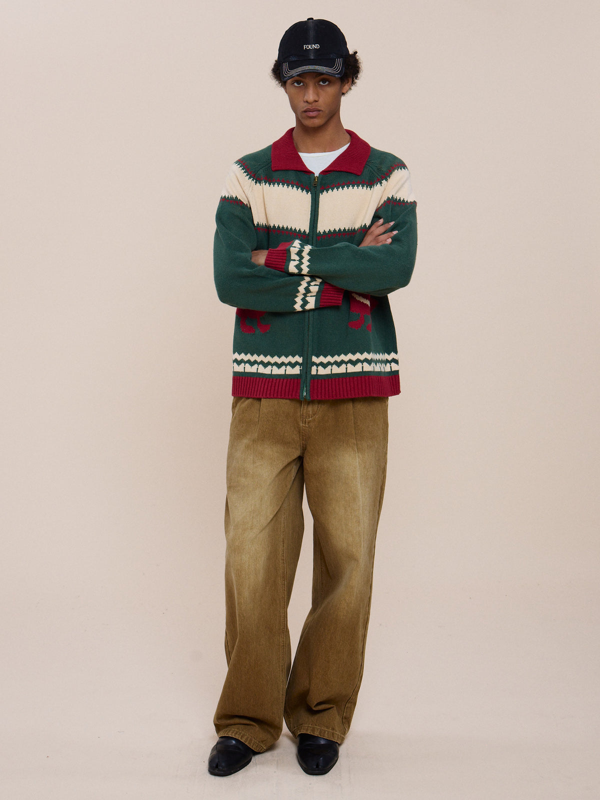 Person standing against a plain background wearing the Isles Sweater by FOUND, a black cap, blue shirt, loose brown pants, and black shoes with arms crossed.