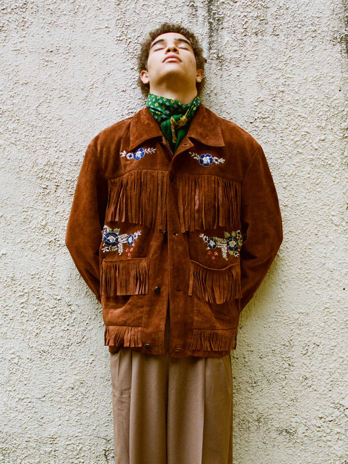 A person stands against a textured wall in the FOUND Hand Embroidered Suede Fringe Jacket, tan pants, and a green patterned scarf, with their head tilted back and eyes closed.