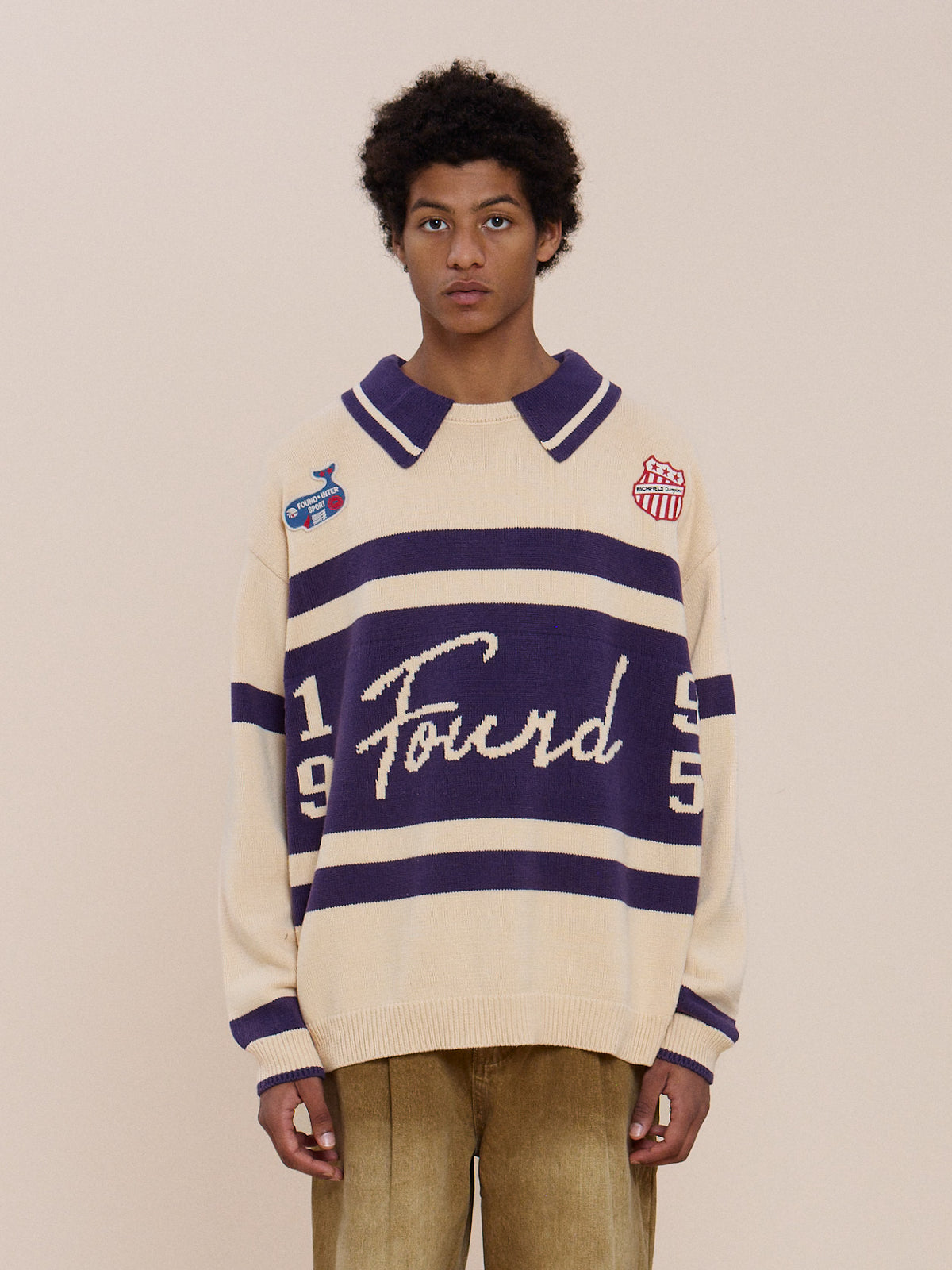 Someone wears the FOUND Knit Jersey with cream and purple stripes, “Found” and “1995” on the front, embroidered patches, and tan pants against a plain beige background.
