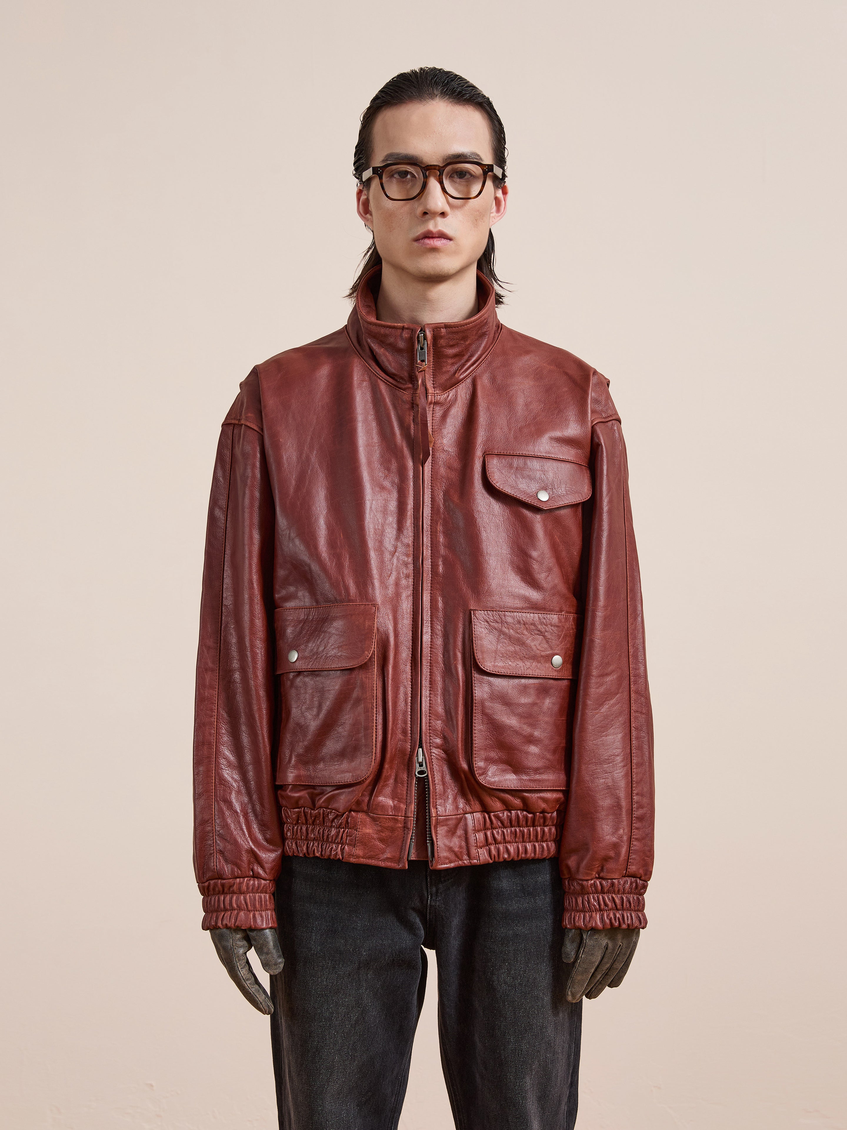 A person wearing glasses, the Found Redwood Leather Bomber Jacket with multiple pockets, and dark pants stands against a plain beige background.