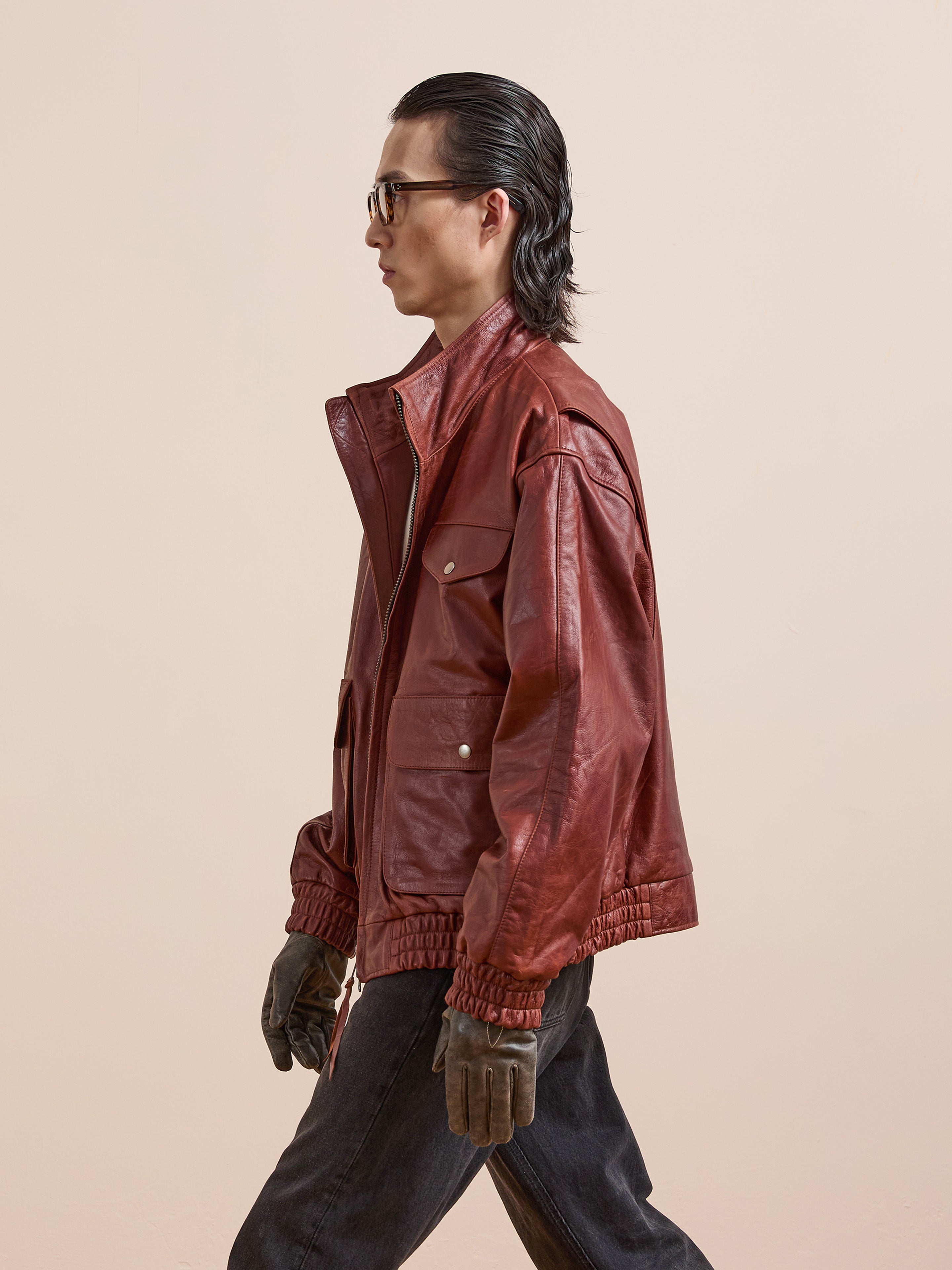 A person in profile wearing the Found Redwood Leather Bomber Jacket, sunglasses, brown gloves, and dark pants stands against a beige backdrop for an effortlessly cool unisex look.