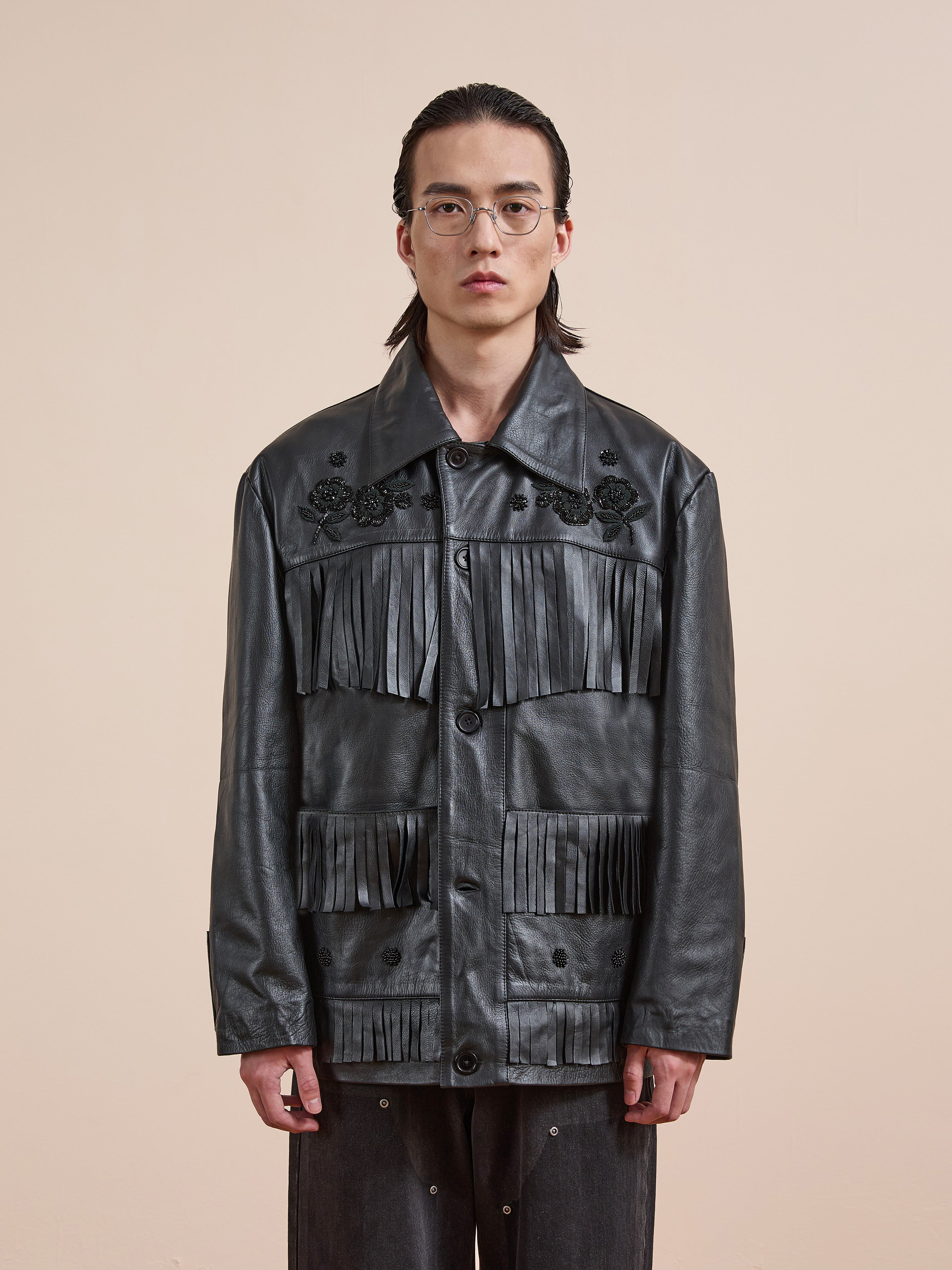 A person with long hair and glasses models the FOUND Hand Embroidered Leather Fringe Jacket, featuring floral details, against a plain beige background.