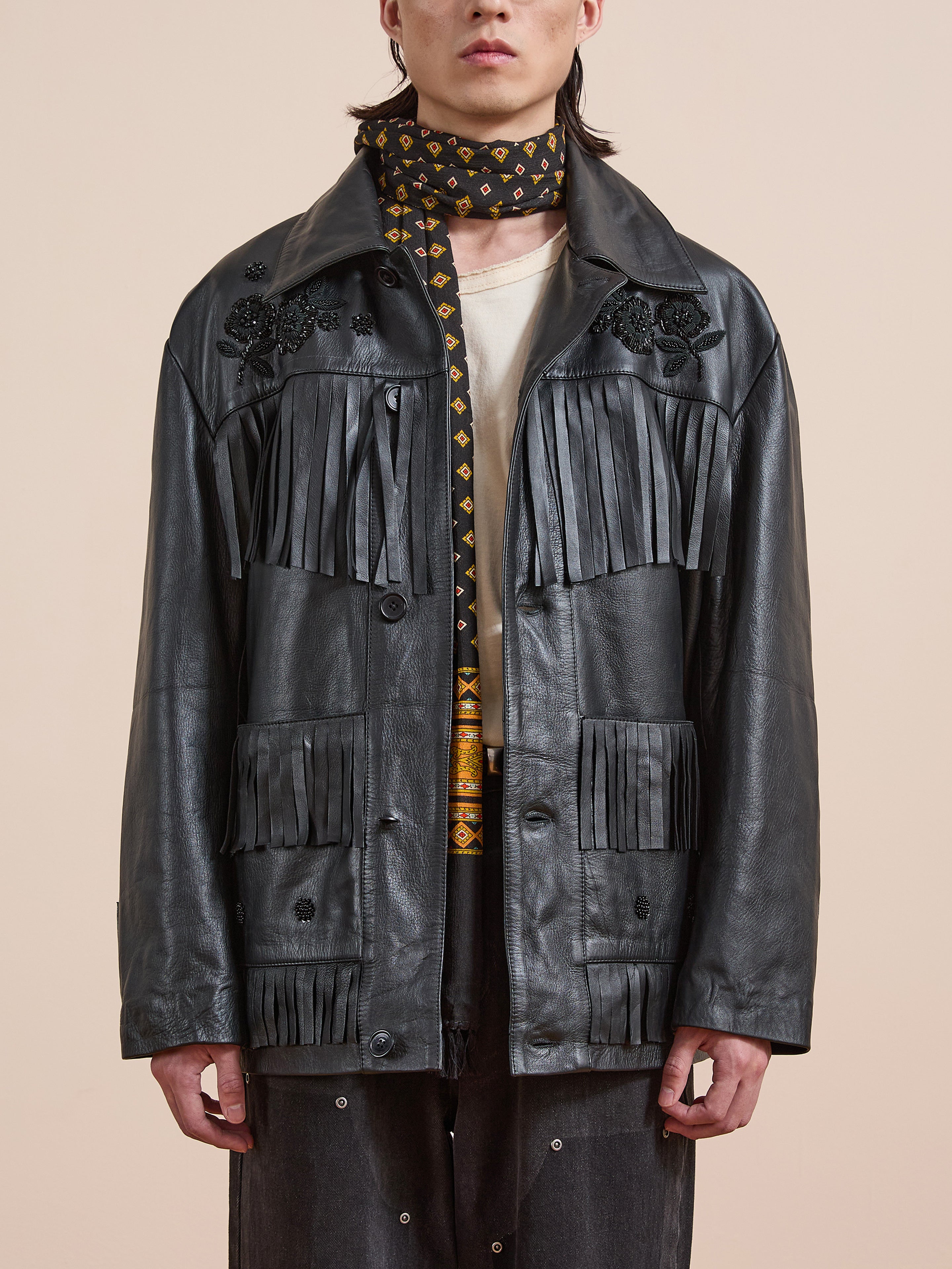 Someone wears the FOUND Hand Embroidered Leather Fringe Jacket, a patterned scarf, a beige shirt, and dark pants, standing against a plain background.