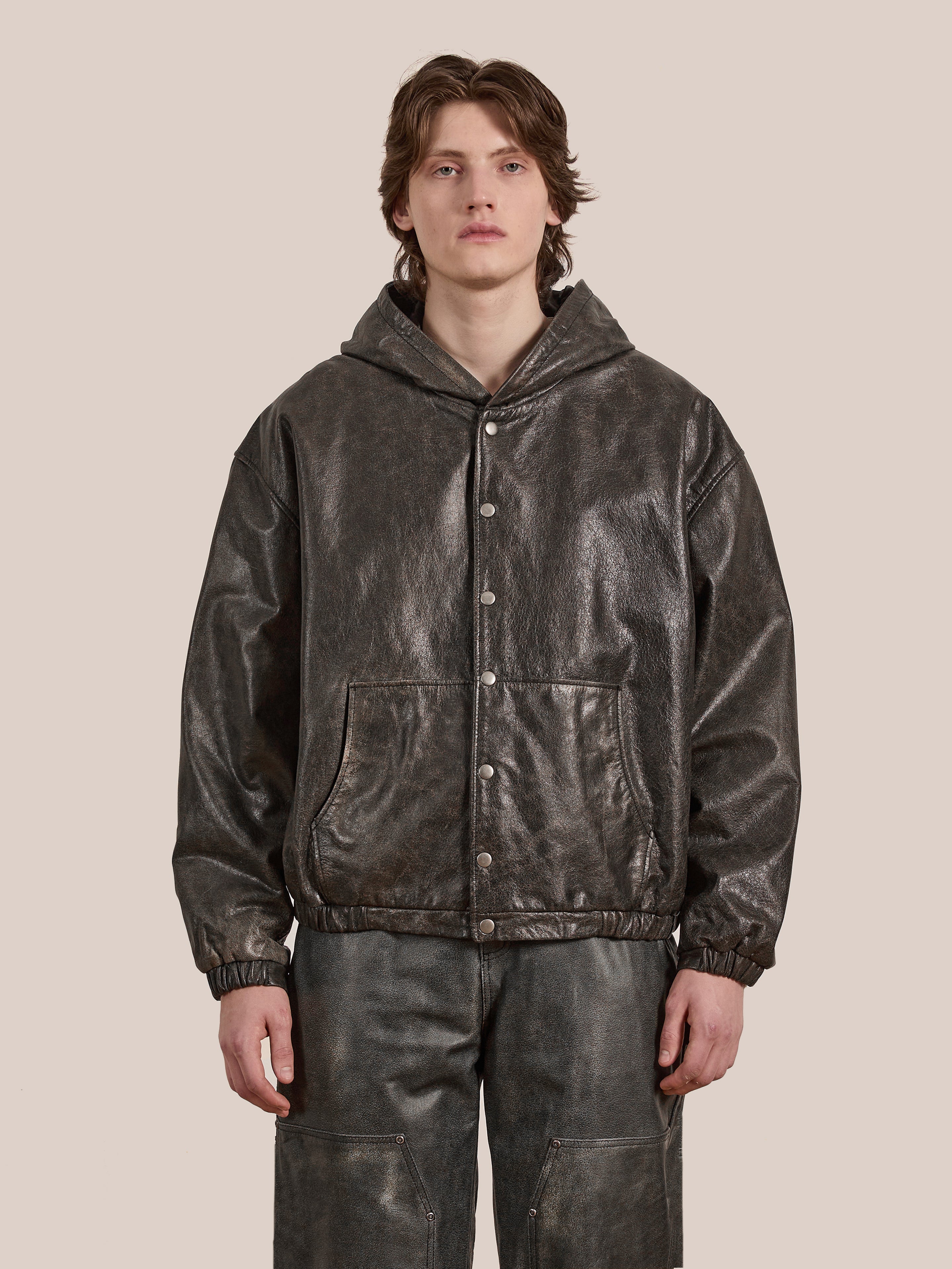 A person wearing a Distressed Genuine Leather Hoodie by Found, featuring a dark metallic pattern and crafted from ethically sourced materials, stands against a plain background.