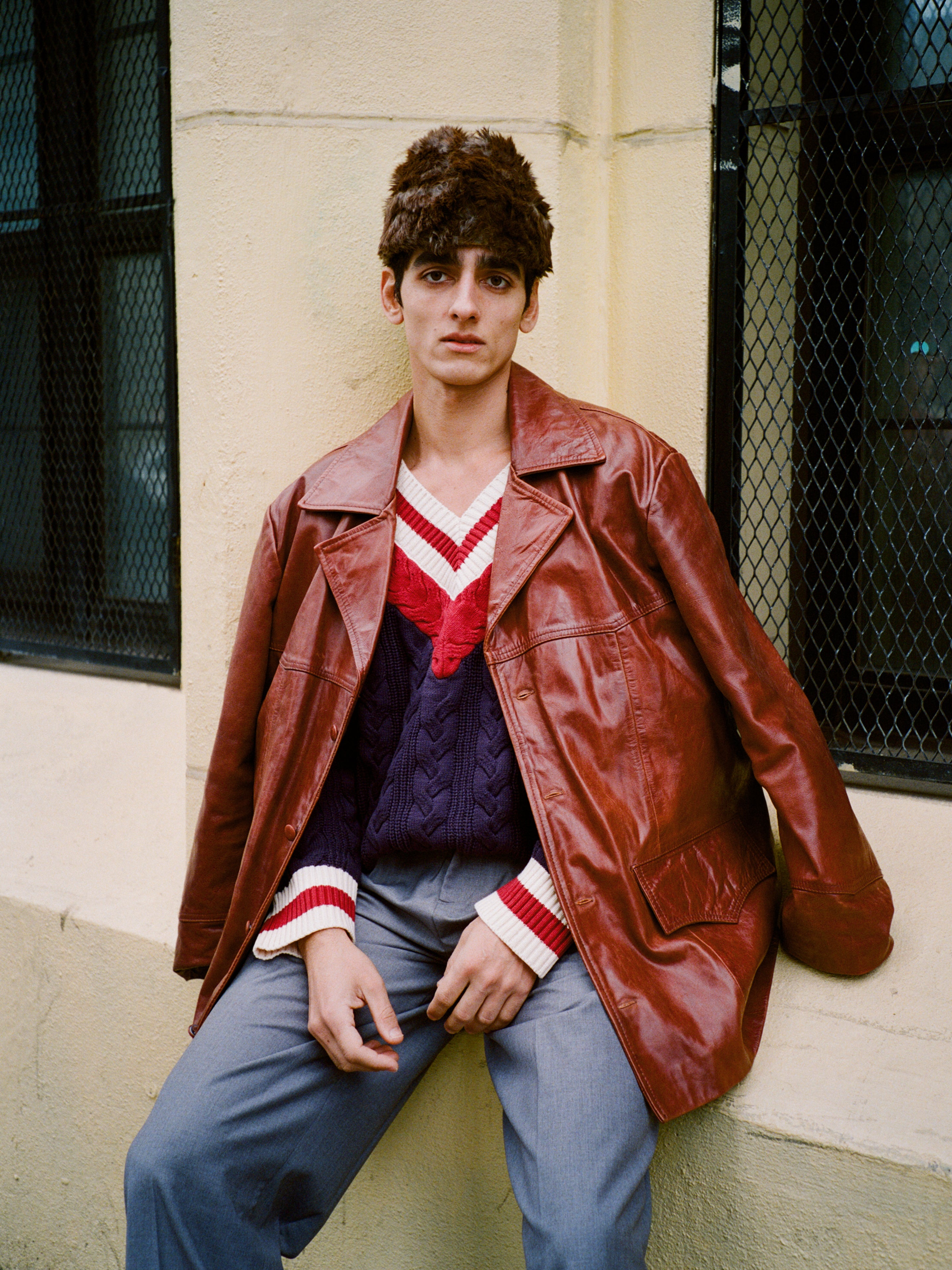 A person in the FOUND Claret Western Oversized Leather Blazer sits against a cream wall and black metal window grilles, styled with a navy sweater featuring red and white trim, gray pants, and a brown fur hat.