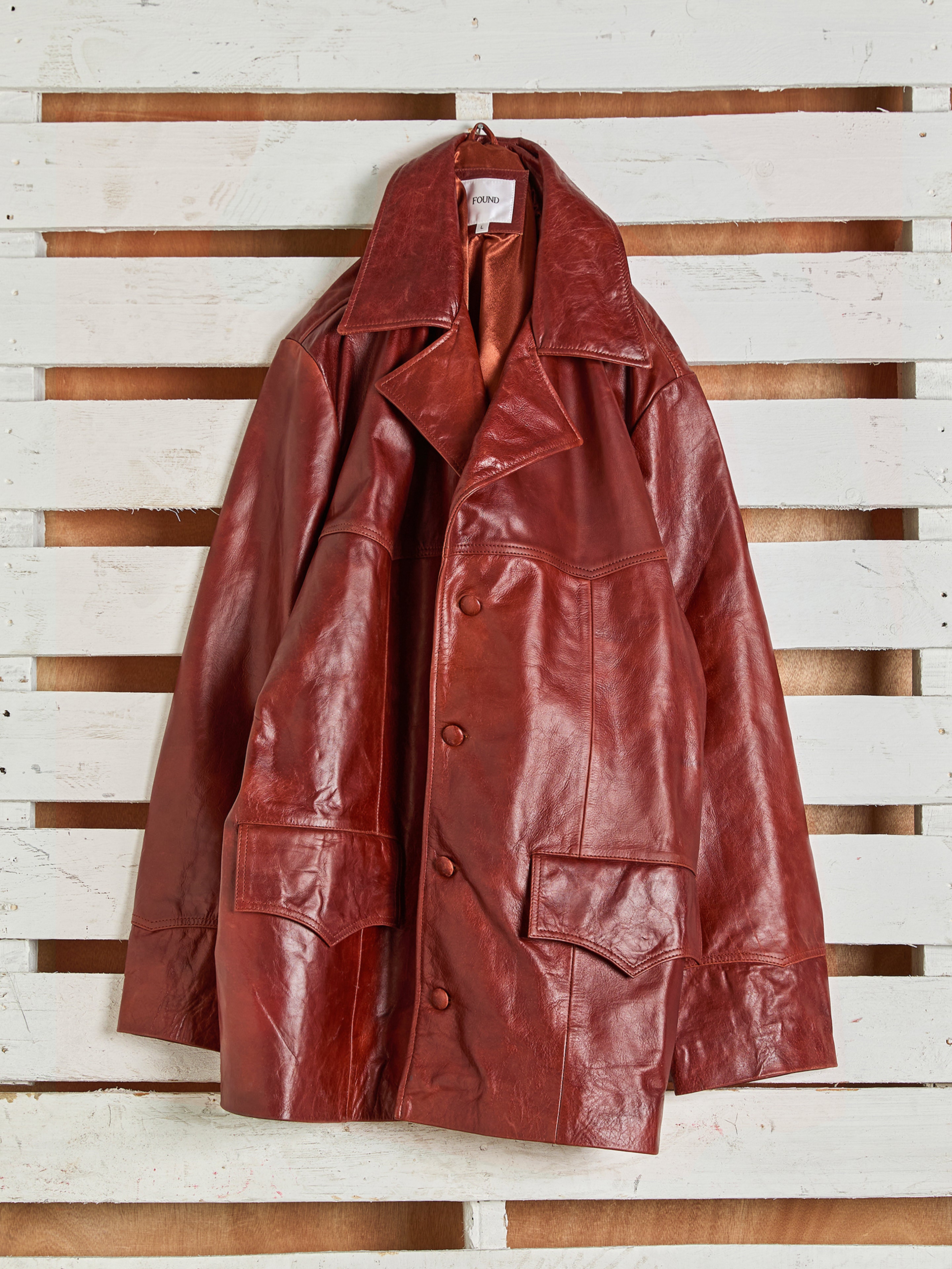 The FOUND Claret Western Oversized Leather Blazer, featuring large front pockets and a wide collar, hangs on a white wooden pallet background.