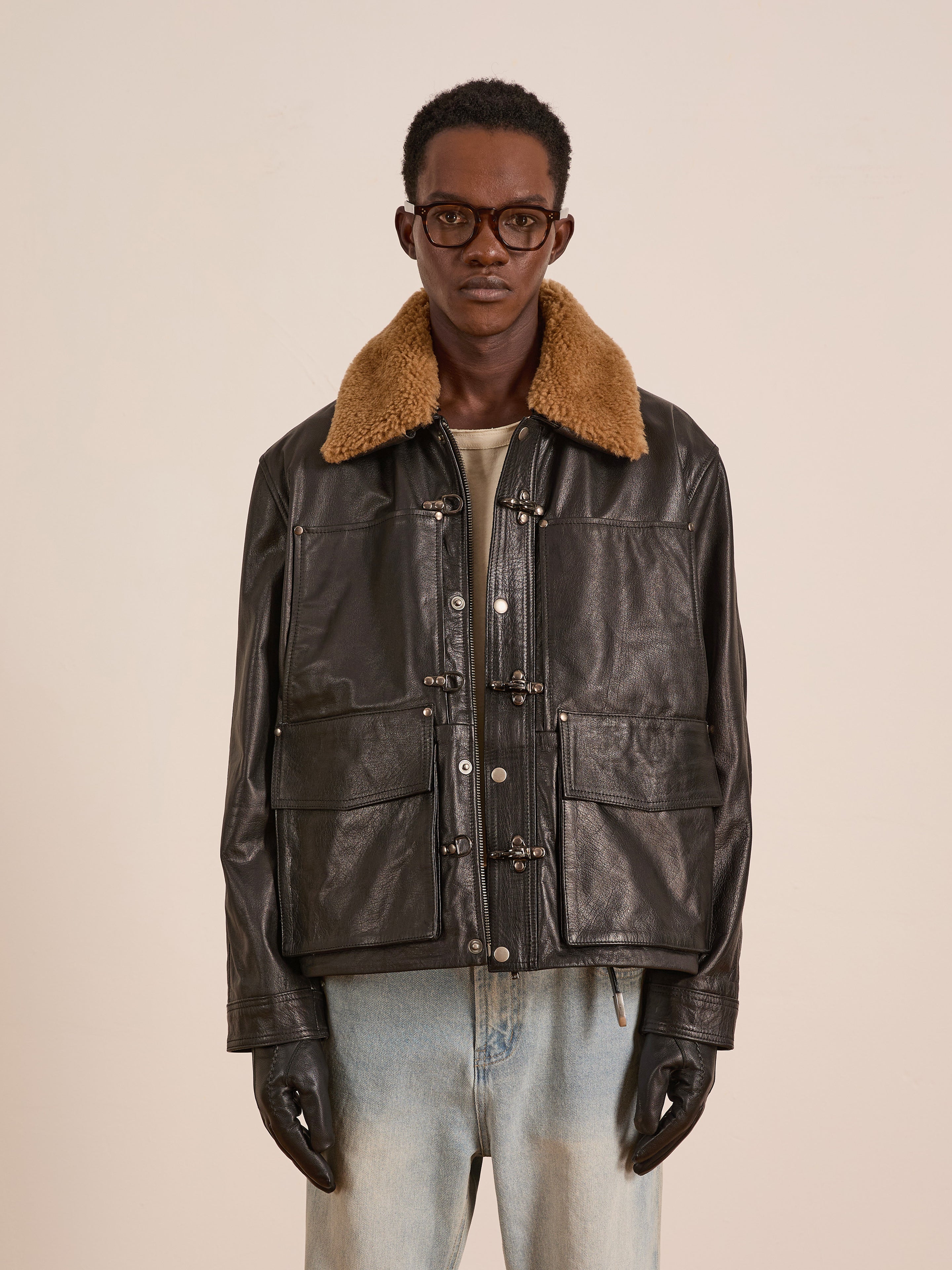 A person wearing glasses, black gloves, a beige shirt, light blue jeans, and the FOUND Sherpa Collar Leather Utility Jacket stands against a plain background.