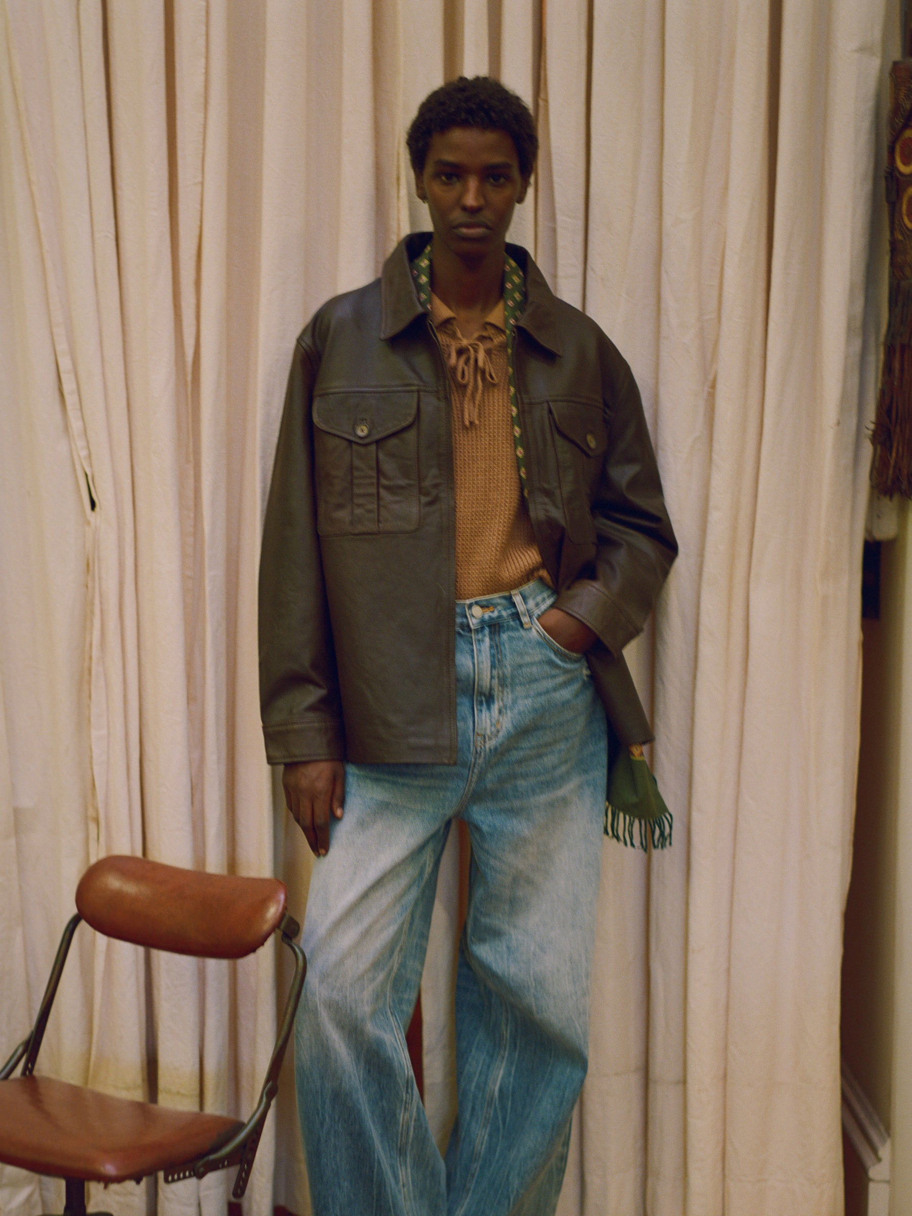 A man in a brown jacket and jeans standing in front of a curtain, wearing a Found Sepia Leather Overshirt.