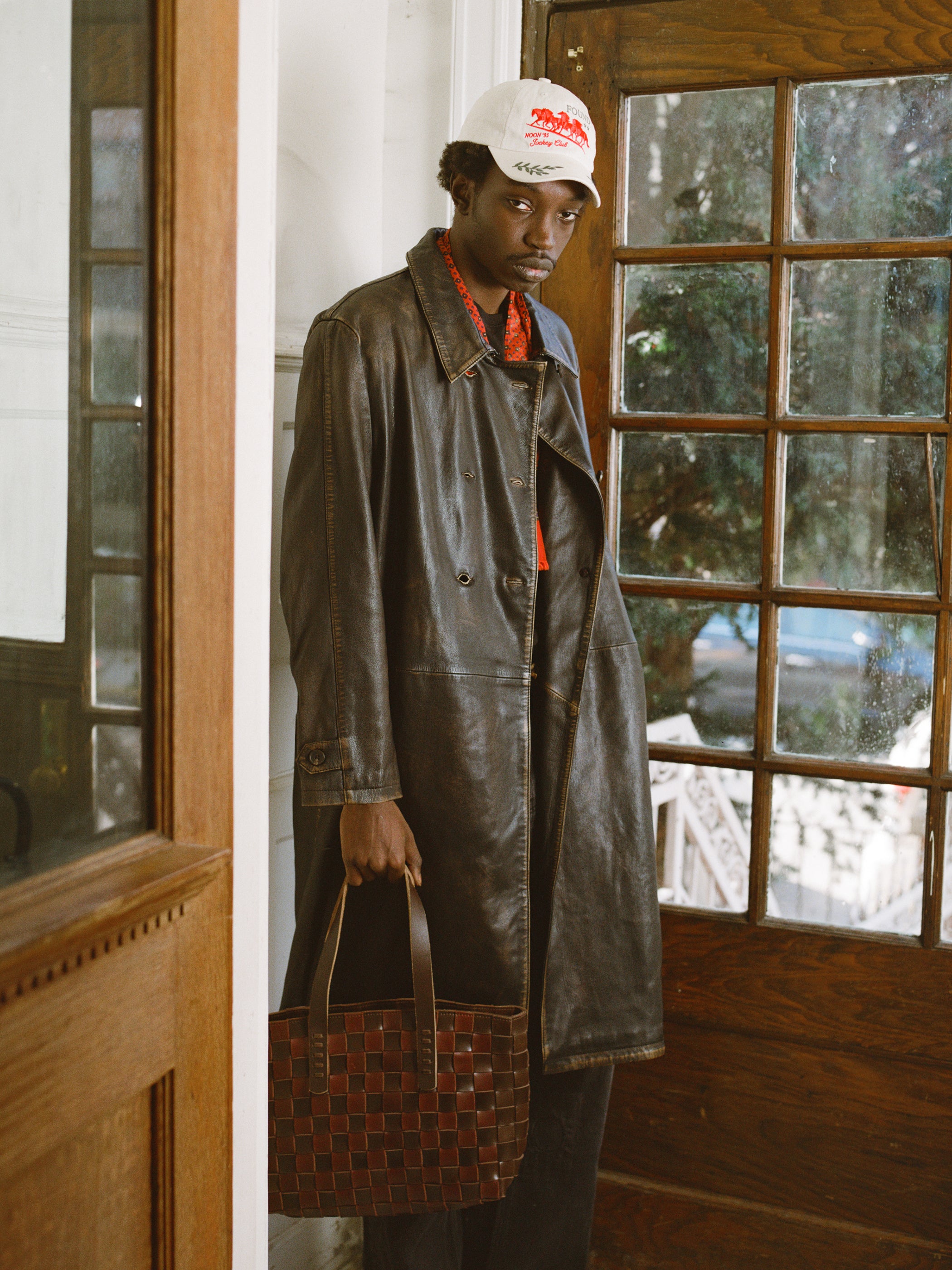 A man stands indoors by a wooden door, wearing FOUND's Distressed Leather Trench Coat, a white cap, and holding a brown woven tote bag.
