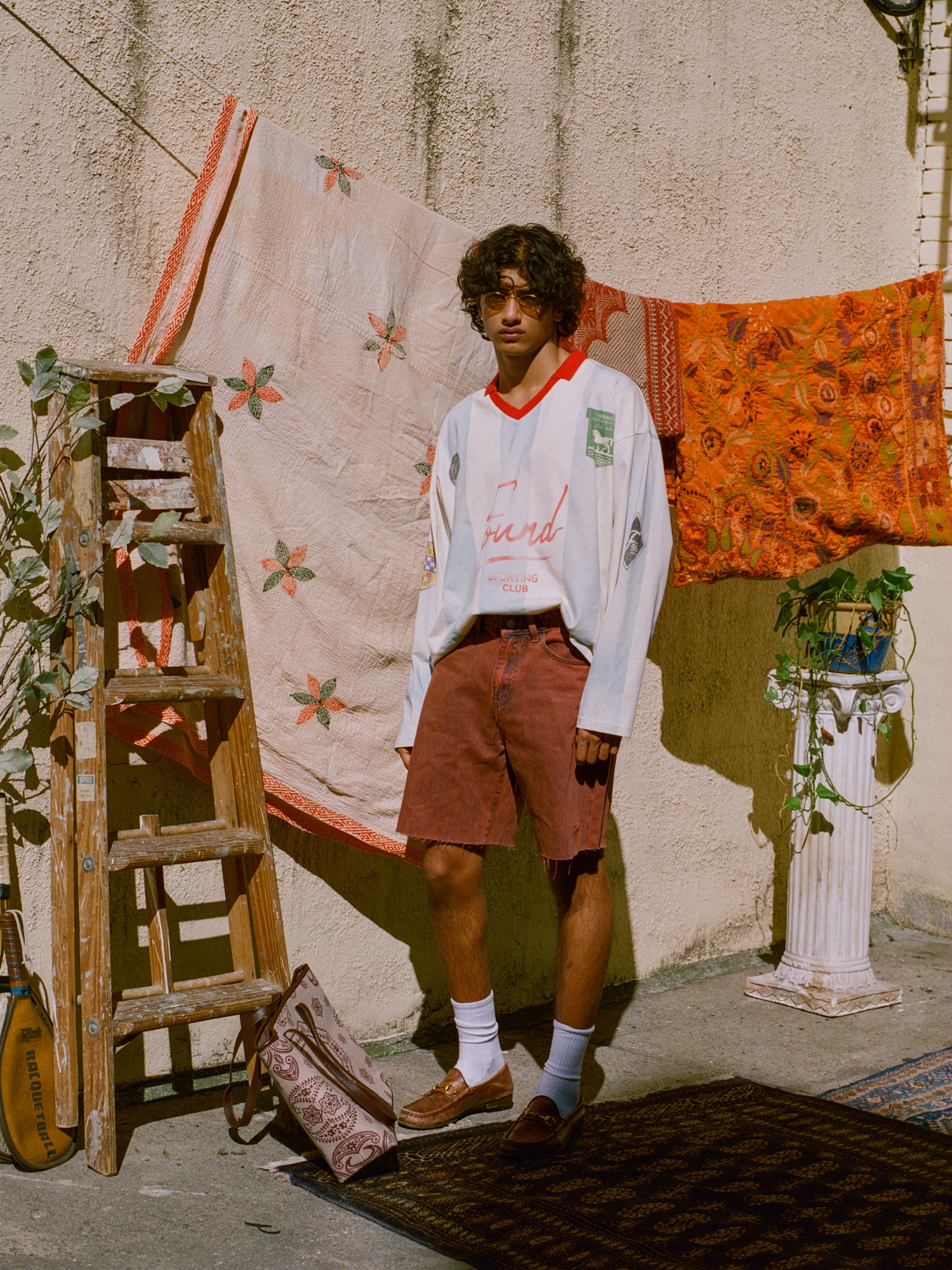 A curly-haired person stands outdoors near a wooden ladder and fabric, wearing the FOUND Striped Sport LS Jersey, red shorts, and loafers. A plant rests on a nearby pedestal.