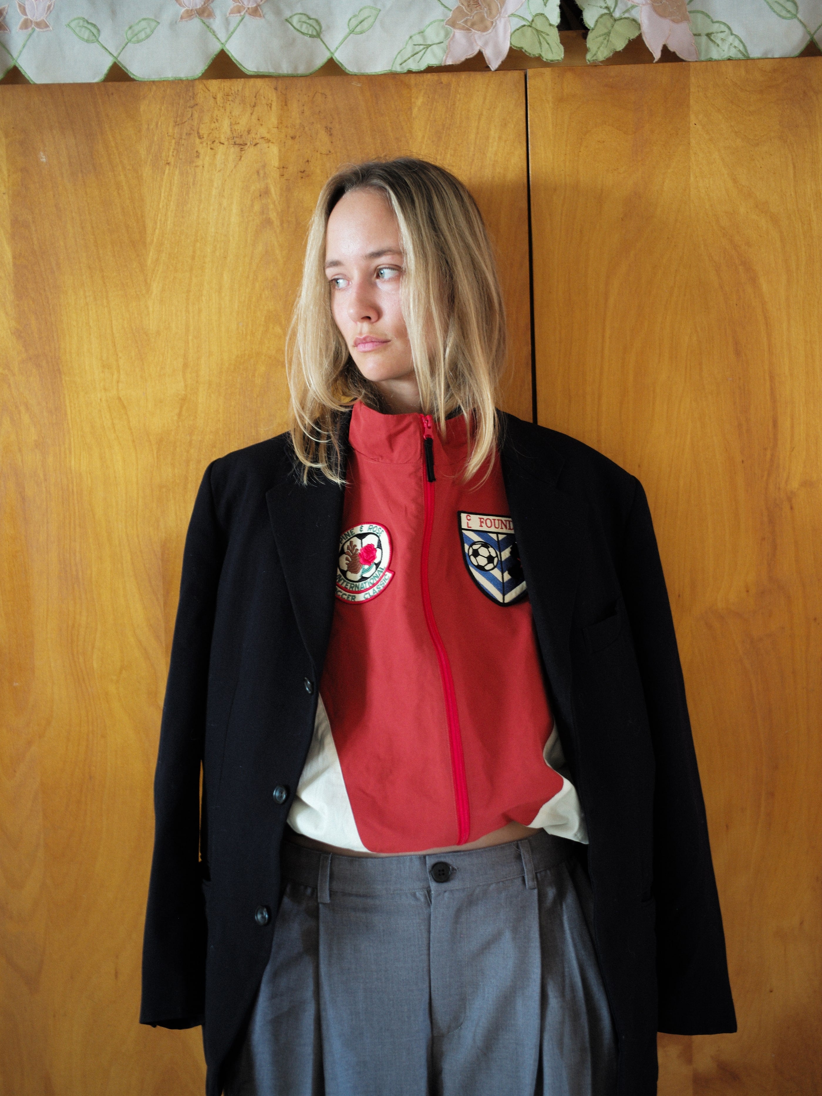 A person with long blond hair dons a red Pine and Rose Track Jacket by FOUND, effortlessly uniting unisex style with sophistication over a black blazer and gray pants. They stand against a wooden backdrop, gazing thoughtfully to the left.
