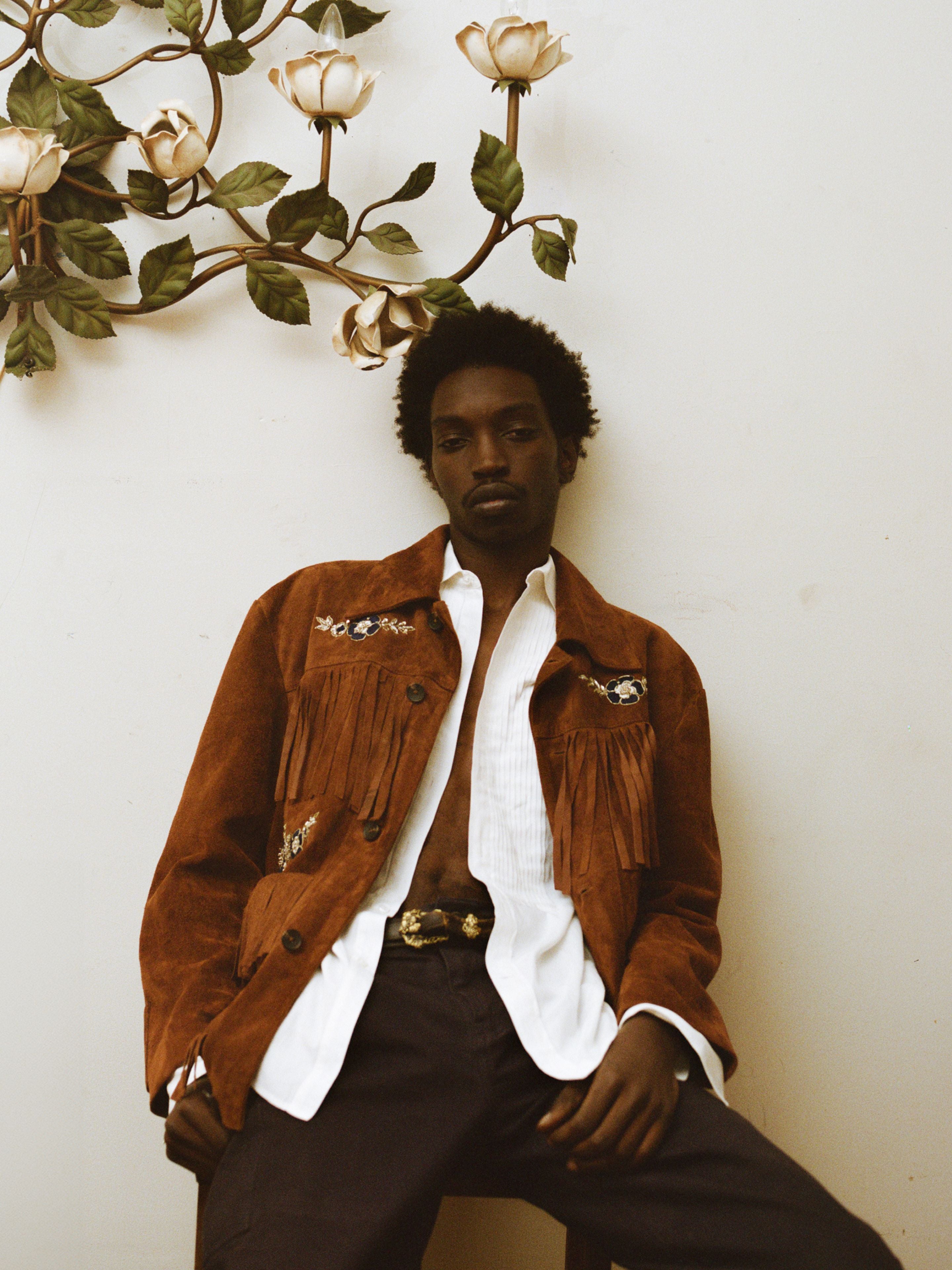 A man in the FOUND Hand Embroidered Suede Fringe Jacket, paired with a white shirt and dark pants, sits against a wall adorned with a floral light fixture.