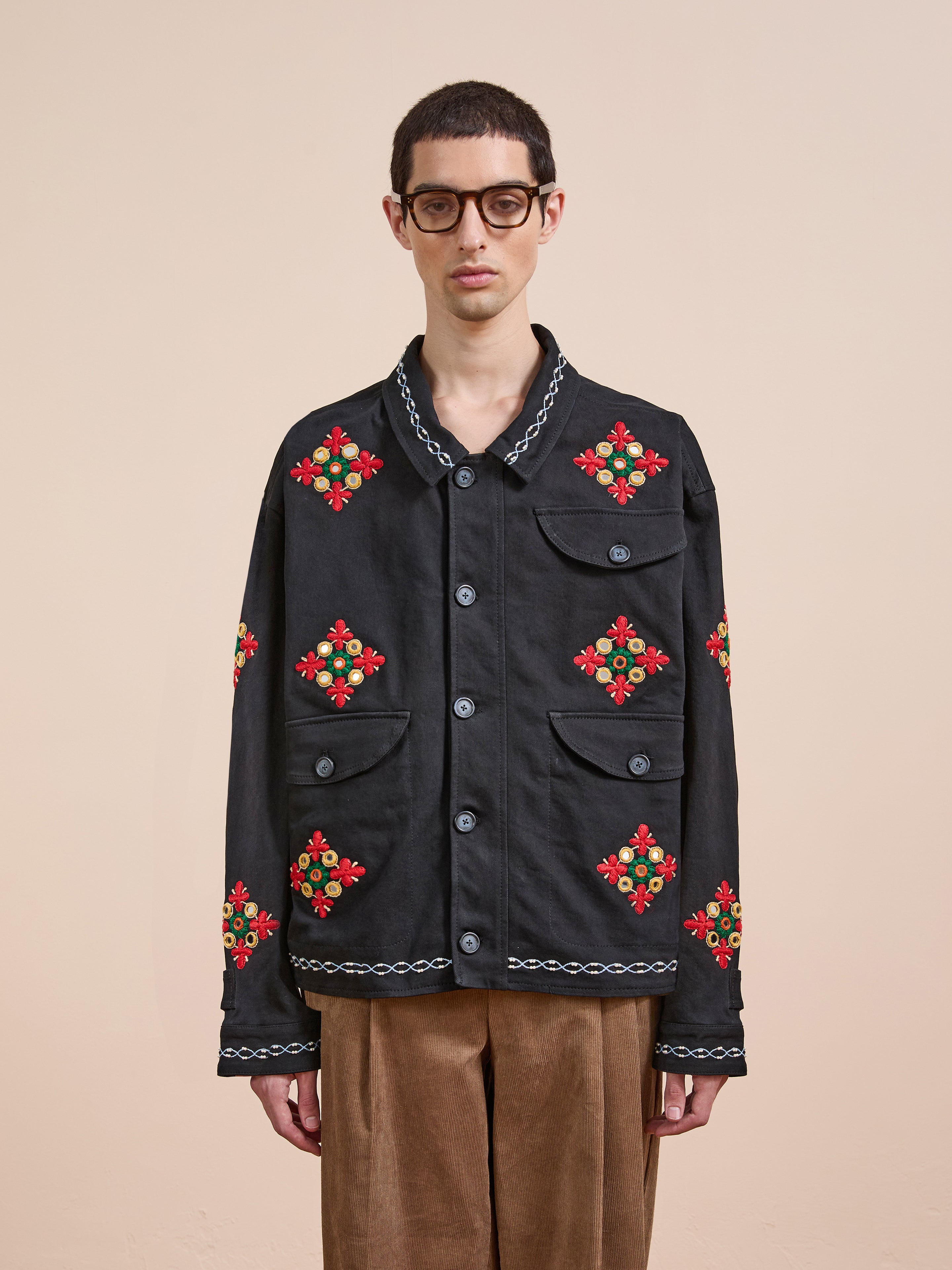 A person wearing glasses and the Found Mirror Ornament Hand Embroidered Work Jacket, featuring red and green Aari and Sheeshakari floral designs on black cotton twill, stands against a plain beige background.