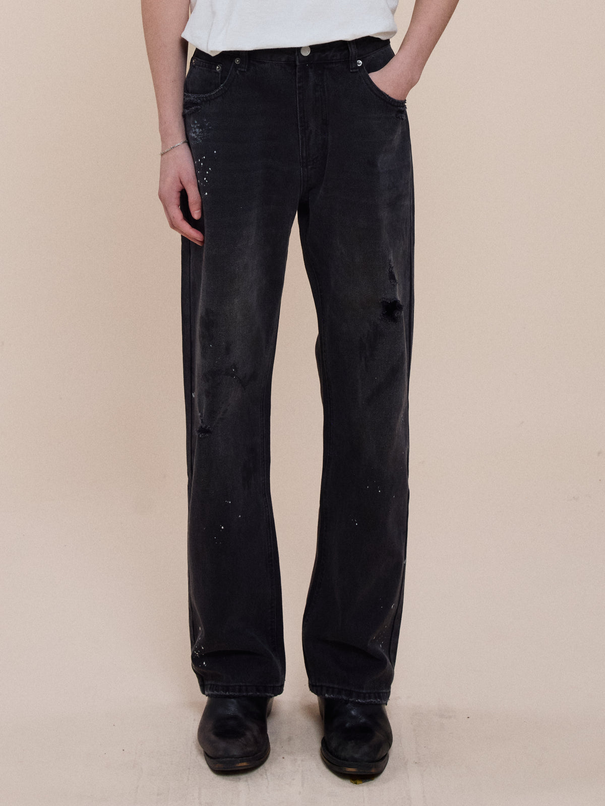 Person wearing Monet Paint Jeans by FOUND, a white t-shirt, and black shoes, standing with one hand in their pocket against a plain beige background.