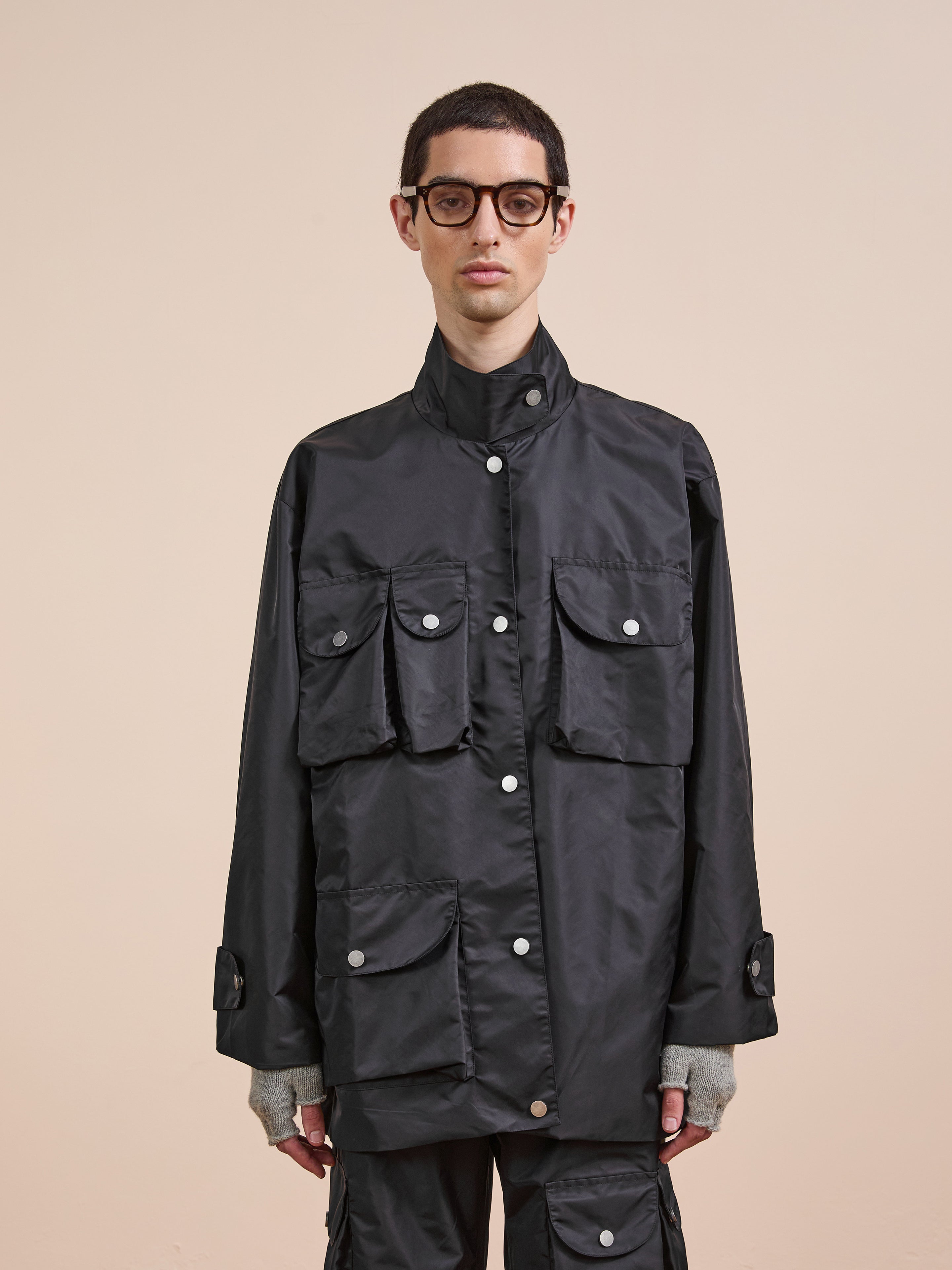 A person wearing glasses and the FOUND Cargo Nylon Jacket, a black unisex piece with multiple pockets, stands against a plain beige background.
