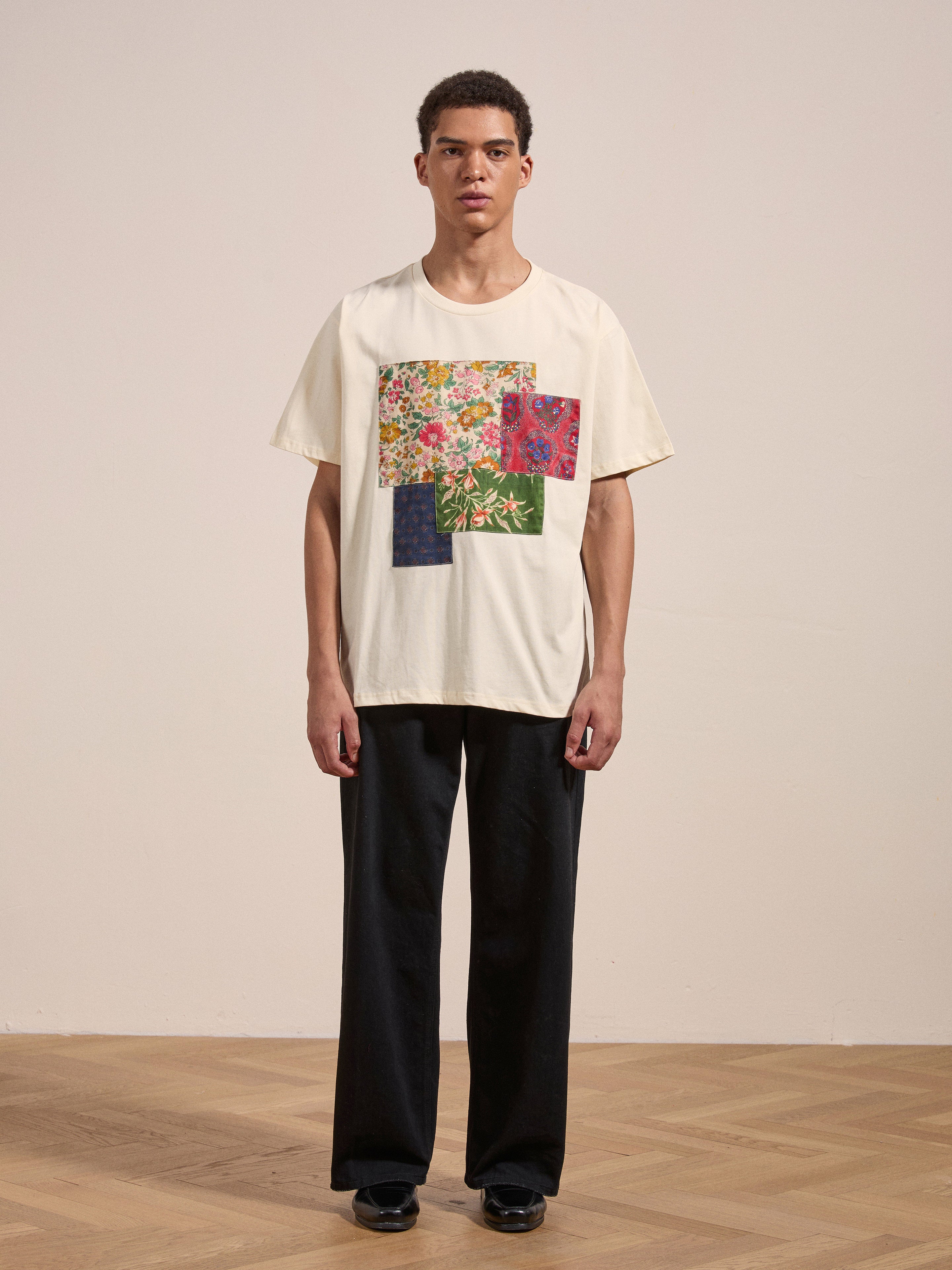 A young man stands on a wooden floor against a plain background, wearing FOUND's Patchwork Tee and black pants.