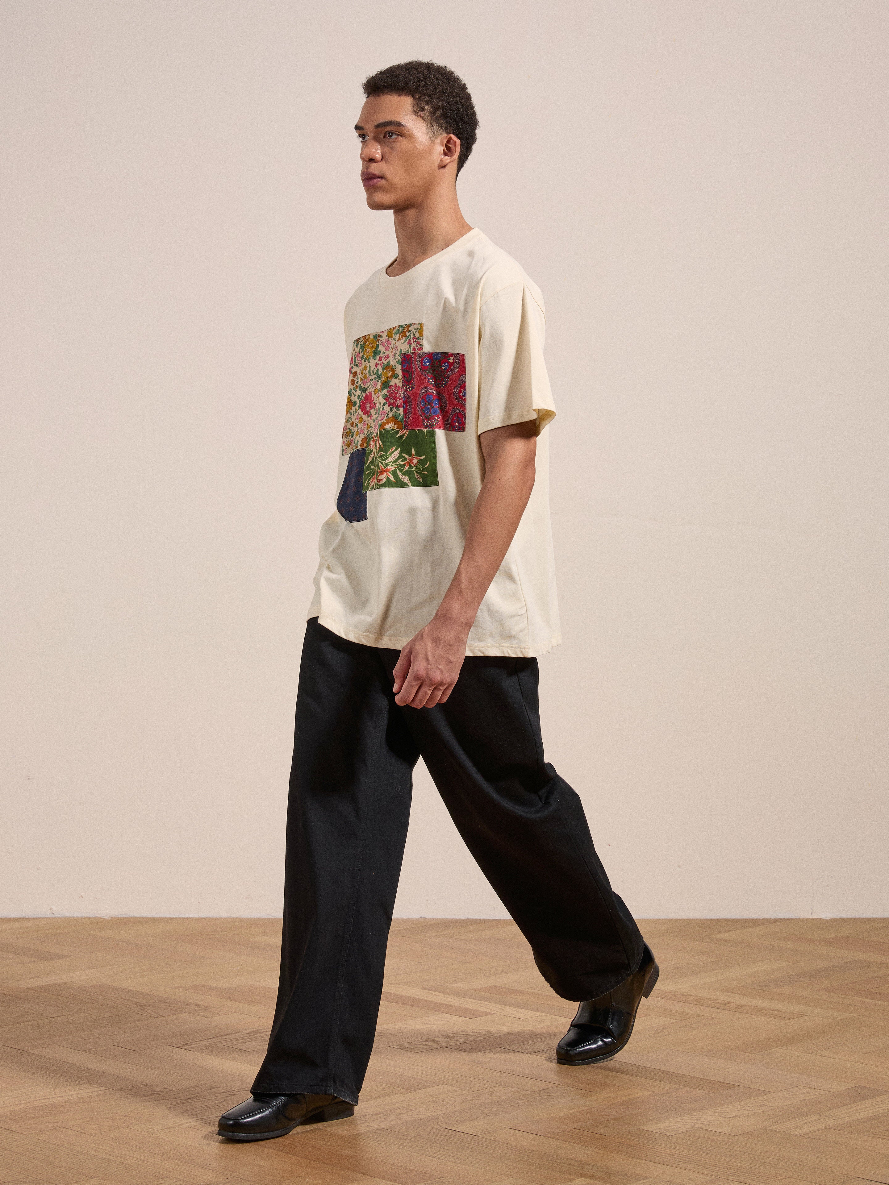 A person wearing a beige Patchwork Tee with colorful patterns from FOUND and black pants walks across a wooden floor in a minimalistic indoor setting.