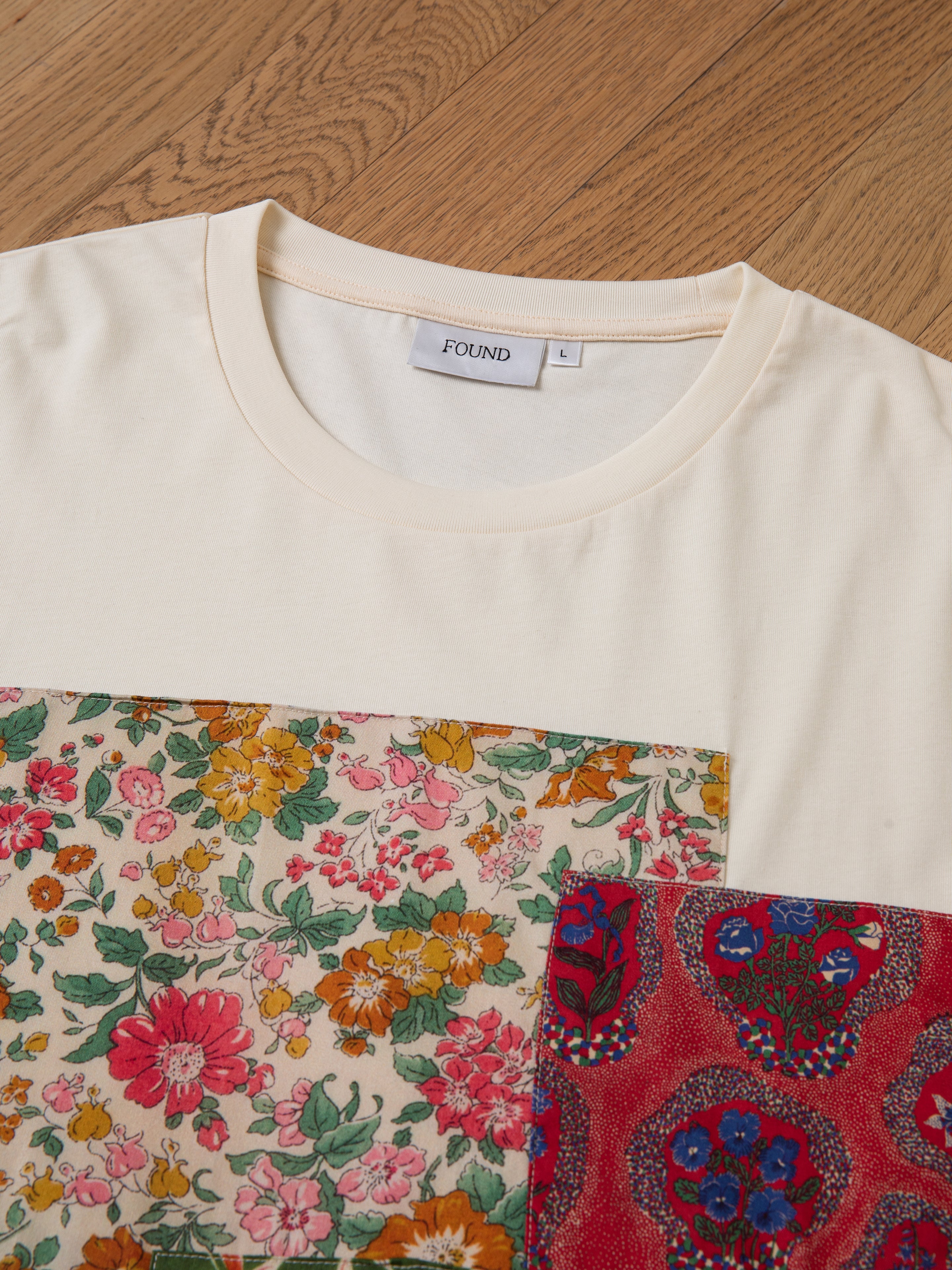 A close-up of a beige Patchwork Tee on a wooden floor, showcasing a floral patch on the front alongside a smaller red patterned patch. The label inside the collar reads "FOUND L.