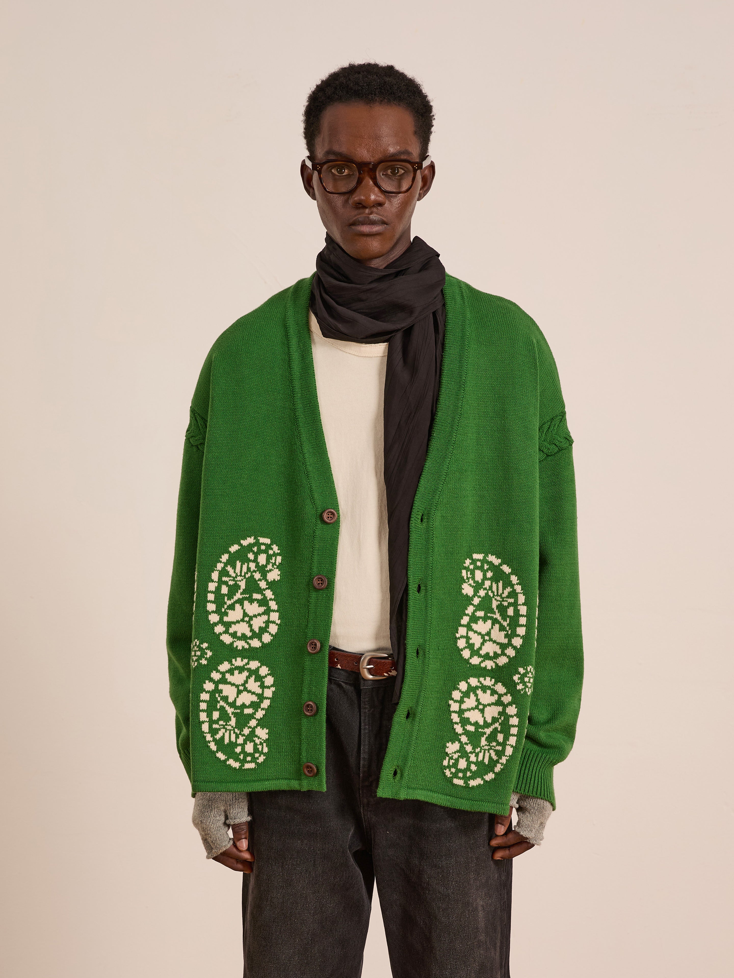 A person wearing glasses, a FOUND Paisley Knit Cardigan with South Asian-inspired patterns, a white shirt, dark jeans, gray fingerless gloves, and a black scarf stands against a plain background.