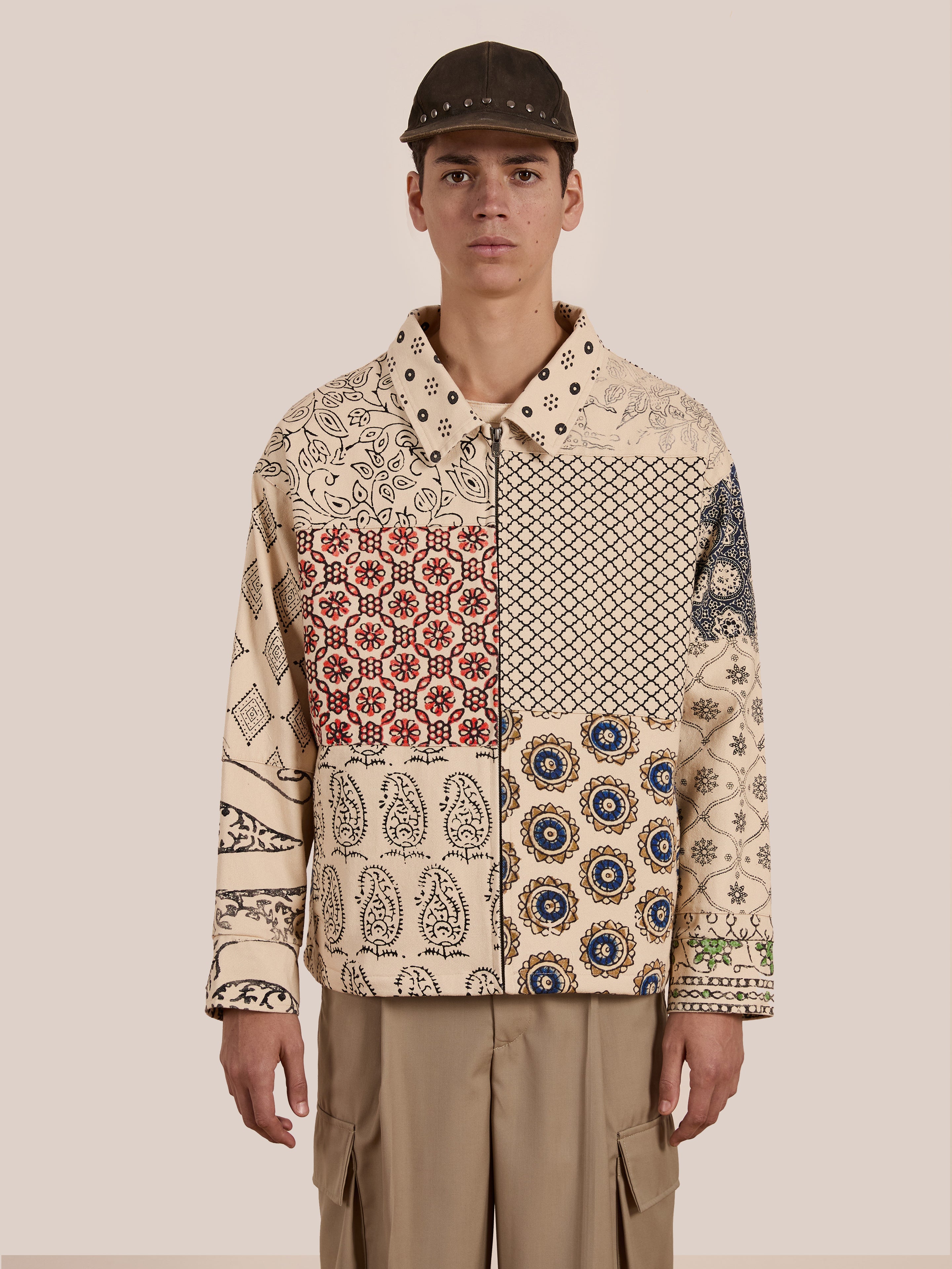 A person stands against a plain background, wearing the Paisley Mosaic Work Jacket by FOUND, made of cotton twill, along with a dark cap.