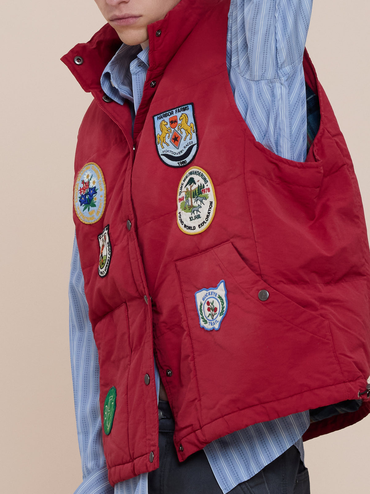Person wearing the FOUND Patch Down Vest with colorful embroidered patches over a blue striped shirt, raising one arm.