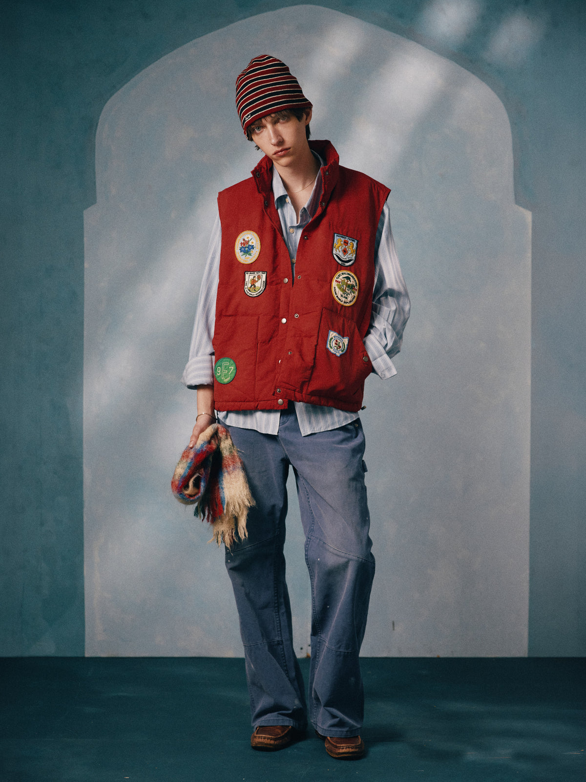 A person wears the FOUND Patch Down Vest, blue shirt, striped beanie, blue pants, and brown shoes, holding a colorful scarf before a blue and white arched backdrop.