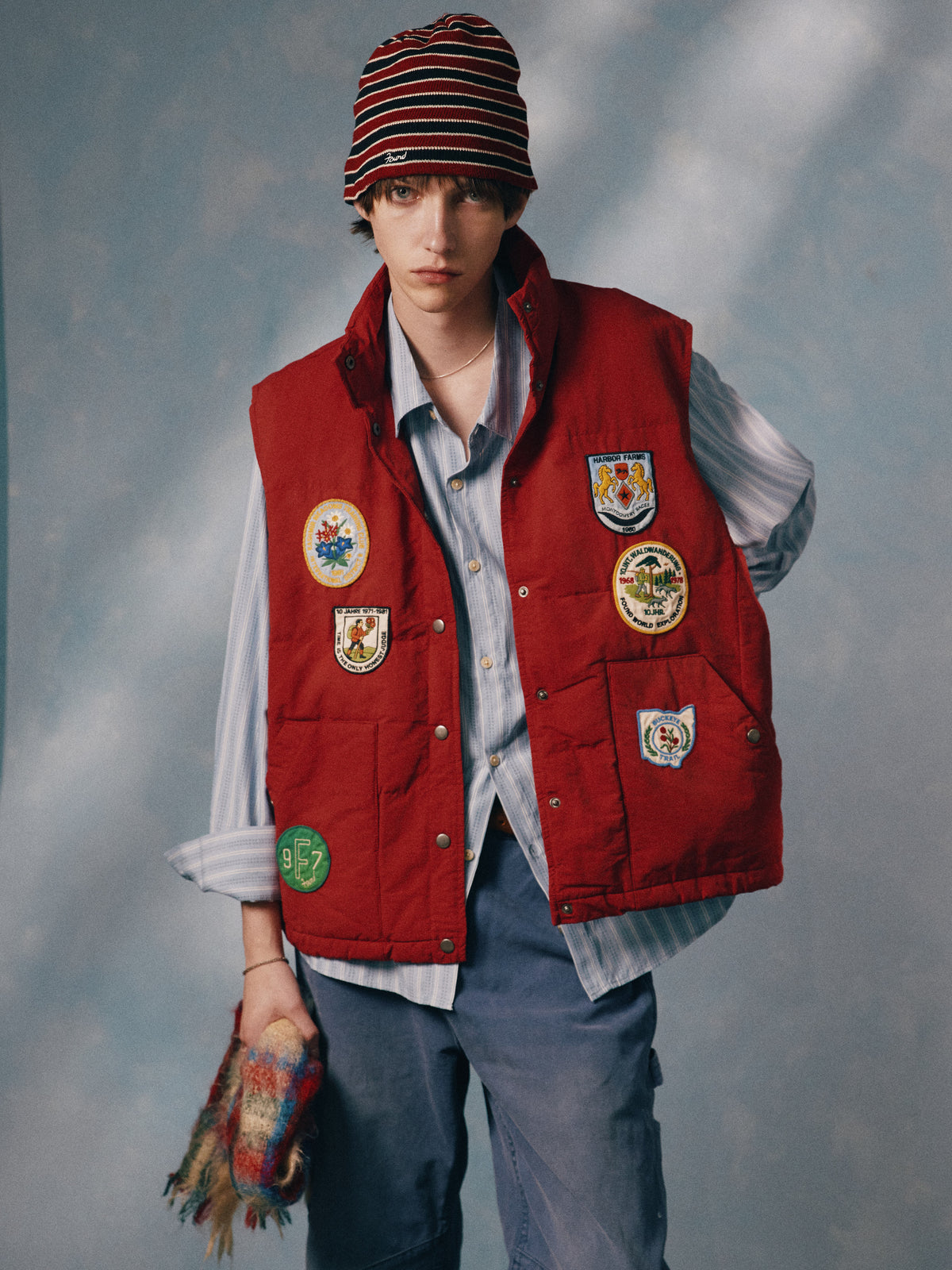 Model wears the FOUND Patch Down Vest with embroidered patches, a striped shirt, blue pants, and beanie, holding a multicolored object against a soft blue background.