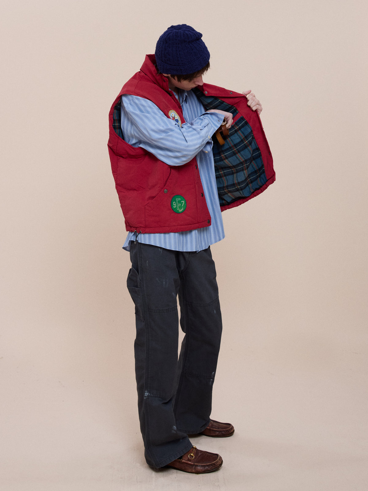 Wearing the FOUND Patch Down Vest, a person in a blue knit hat and striped shirt stands sideways, holding open the vest to show its plaid lining.
