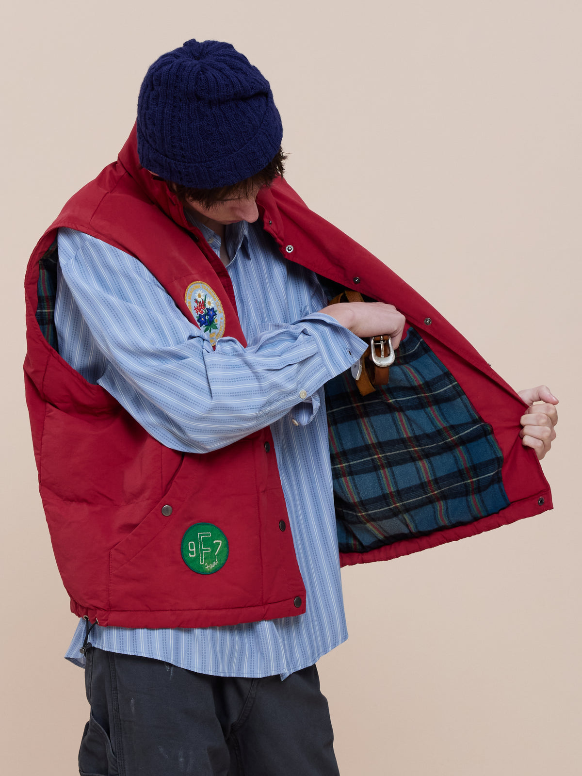 A person in a blue knit hat and striped shirt opens their Patch Down Vest by FOUND to show its blue and brown plaid lining and embroidered patches.