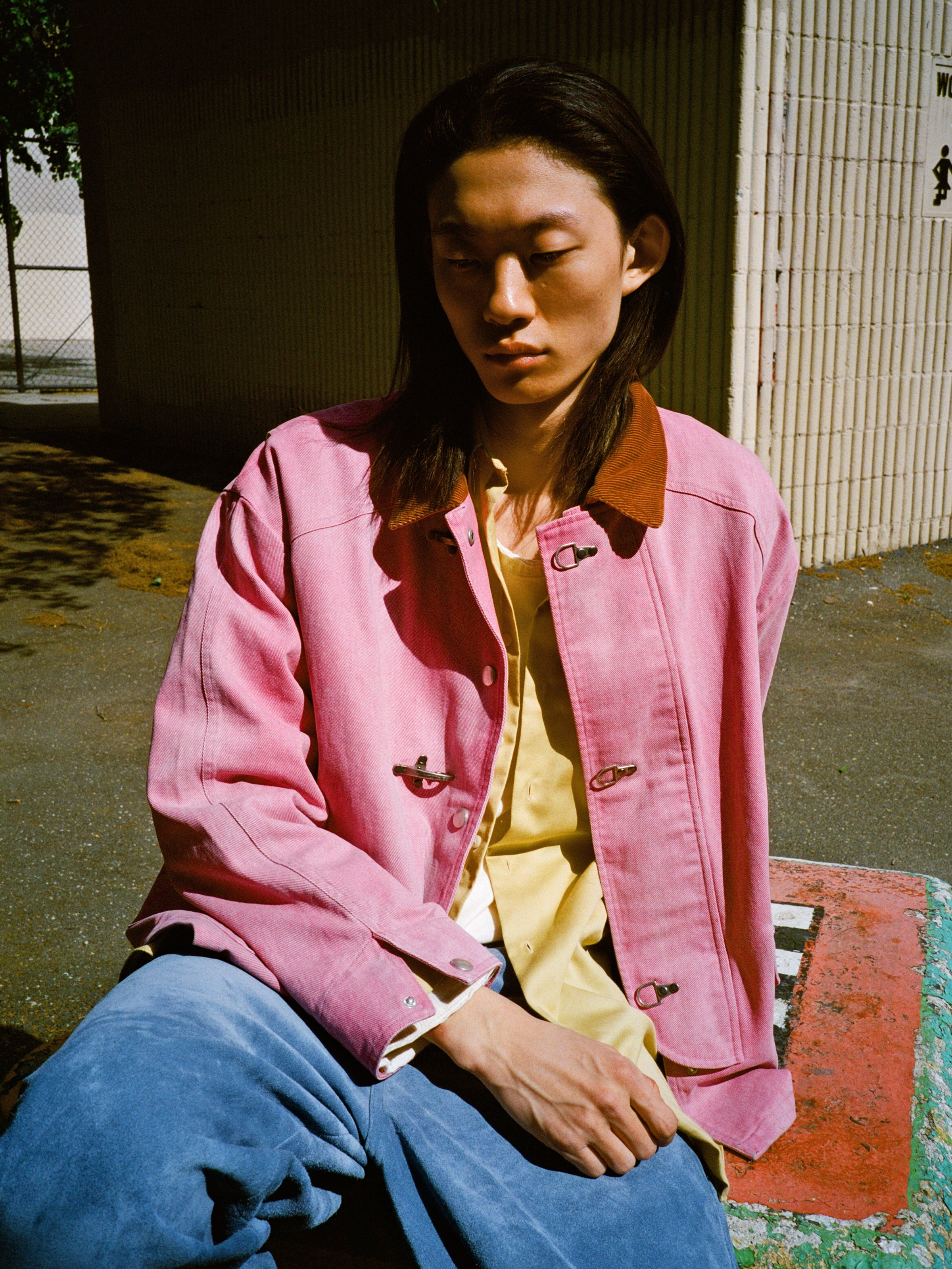 A person with long dark hair sits outdoors, eyes closed as sunlight falls, wearing the Found Washed Fireman Clasp Workshop Jacket paired with a yellow shirt and blue pants.