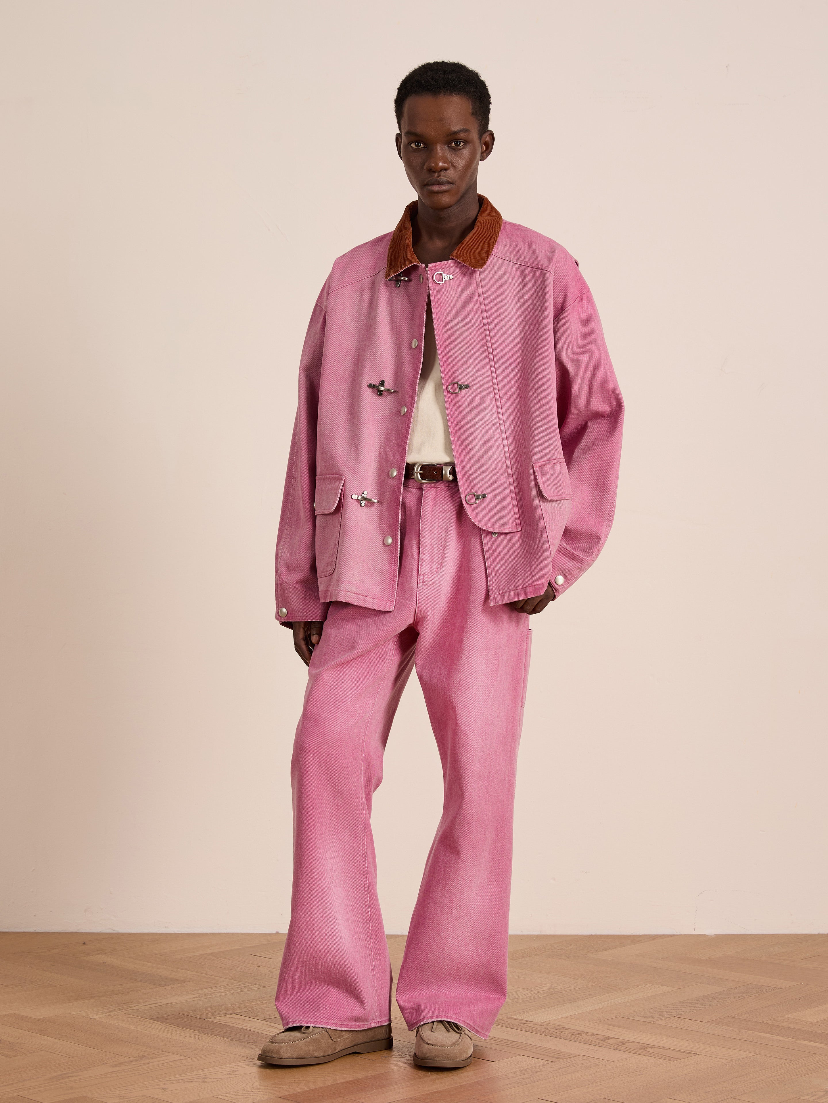 Indoors, a person styles FOUND’s Flare Work Pants in light pink with a matching denim jacket, white shirt, brown-collared jacket, and beige shoes, embodying unisex workwear against a plain background.