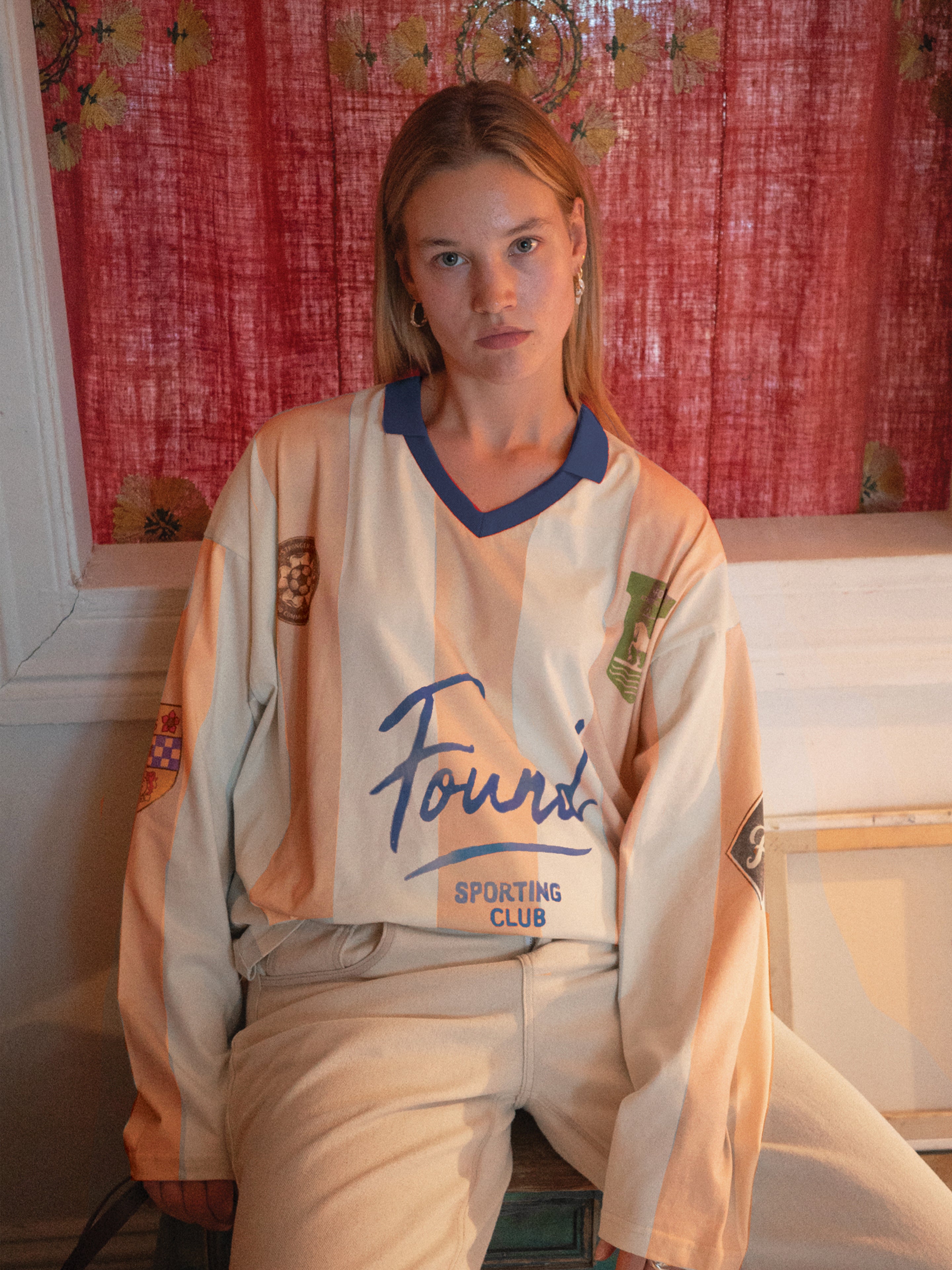 A woman with straight blonde hair sits indoors wearing the FOUND Striped Sport LS Jersey—a unisex, relaxed-fit beige and blue top—paired with light pants, set against a red patterned curtain.