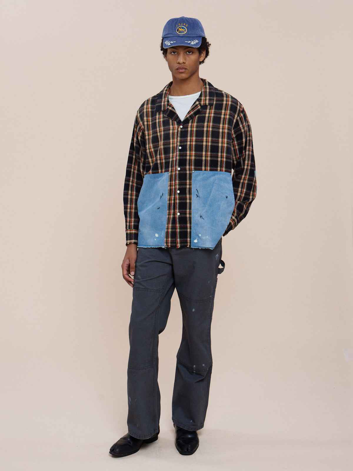Person in a blue cap, FOUND Plaid Denim Shirt, grey pants, and black shoes stands against a plain beige background.