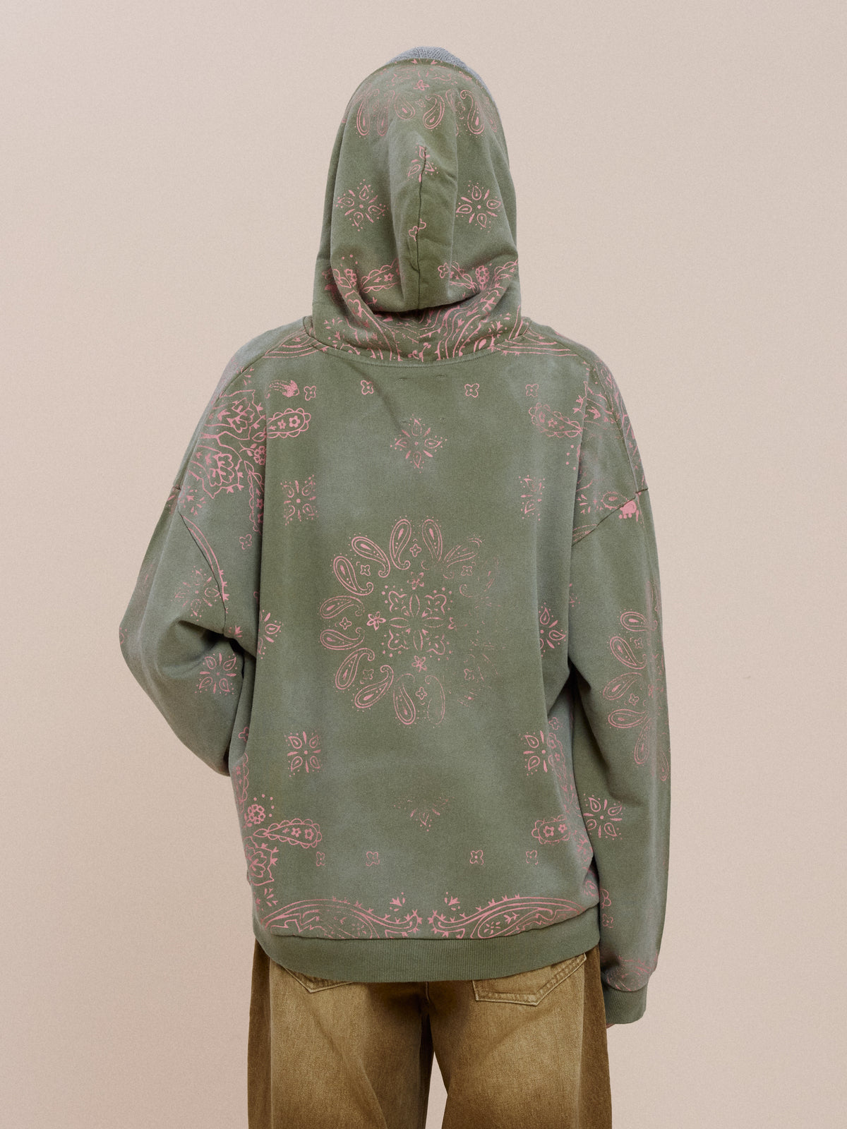 A person wears the FOUND Printed Paisley Hoodie, a green unisex pullover with pink bandana pattern, styled with tan pants on a beige background.