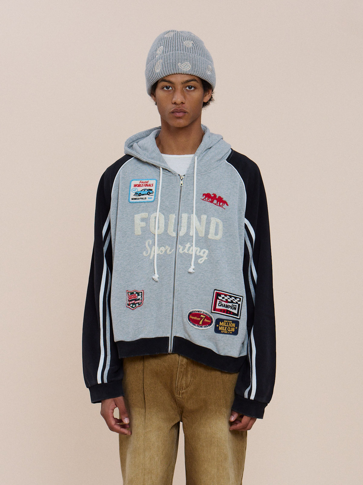 A person in a gray beanie, tan pants, and the FOUND Racer Hoodie with patches stands against a plain light background.