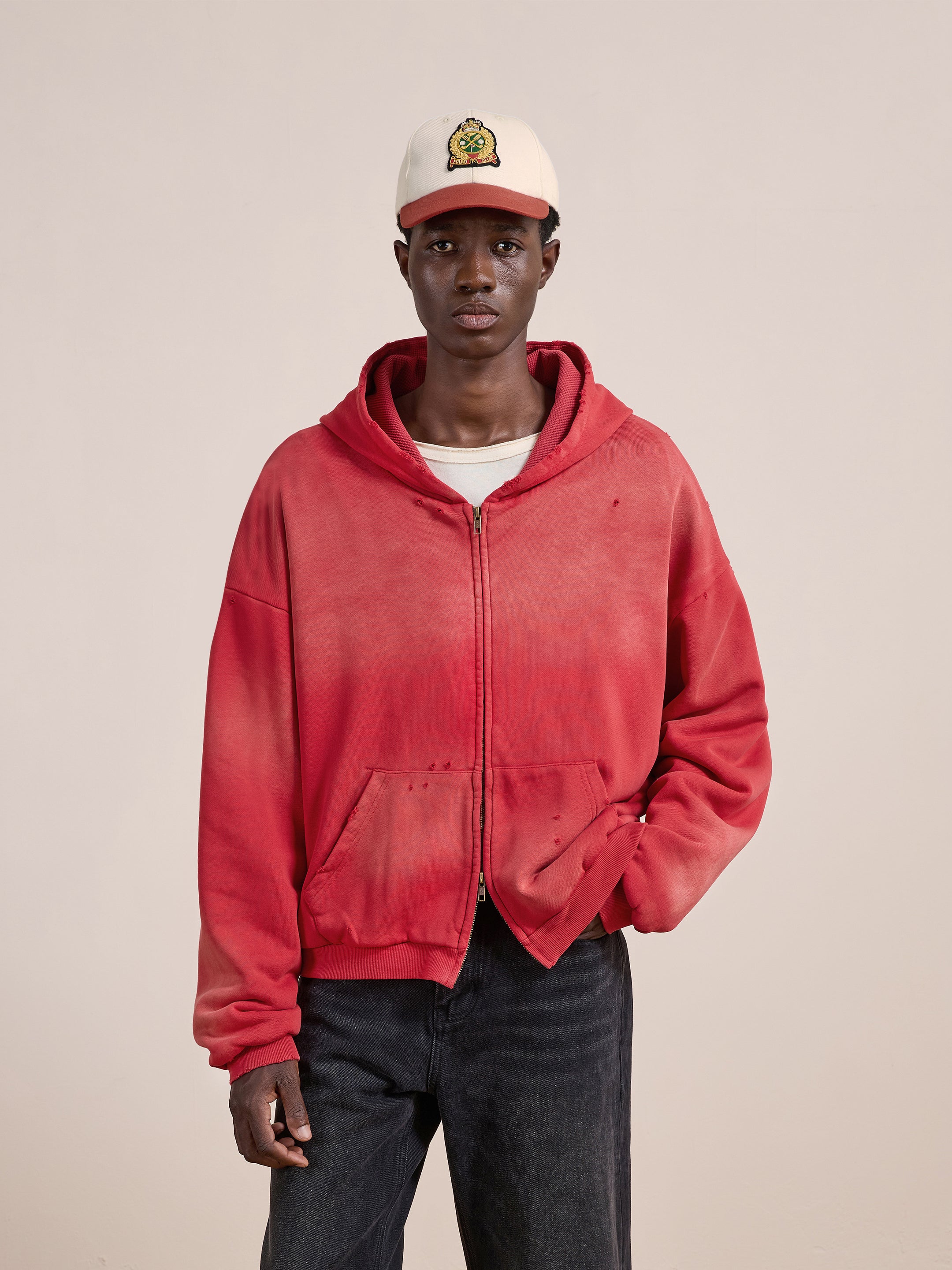 A person wears a FOUND Distressed Thermal Lined Zip Up Hoodie in red, a white t-shirt, black jeans, and a beige cap with a cartoon graphic, standing against a plain light background.