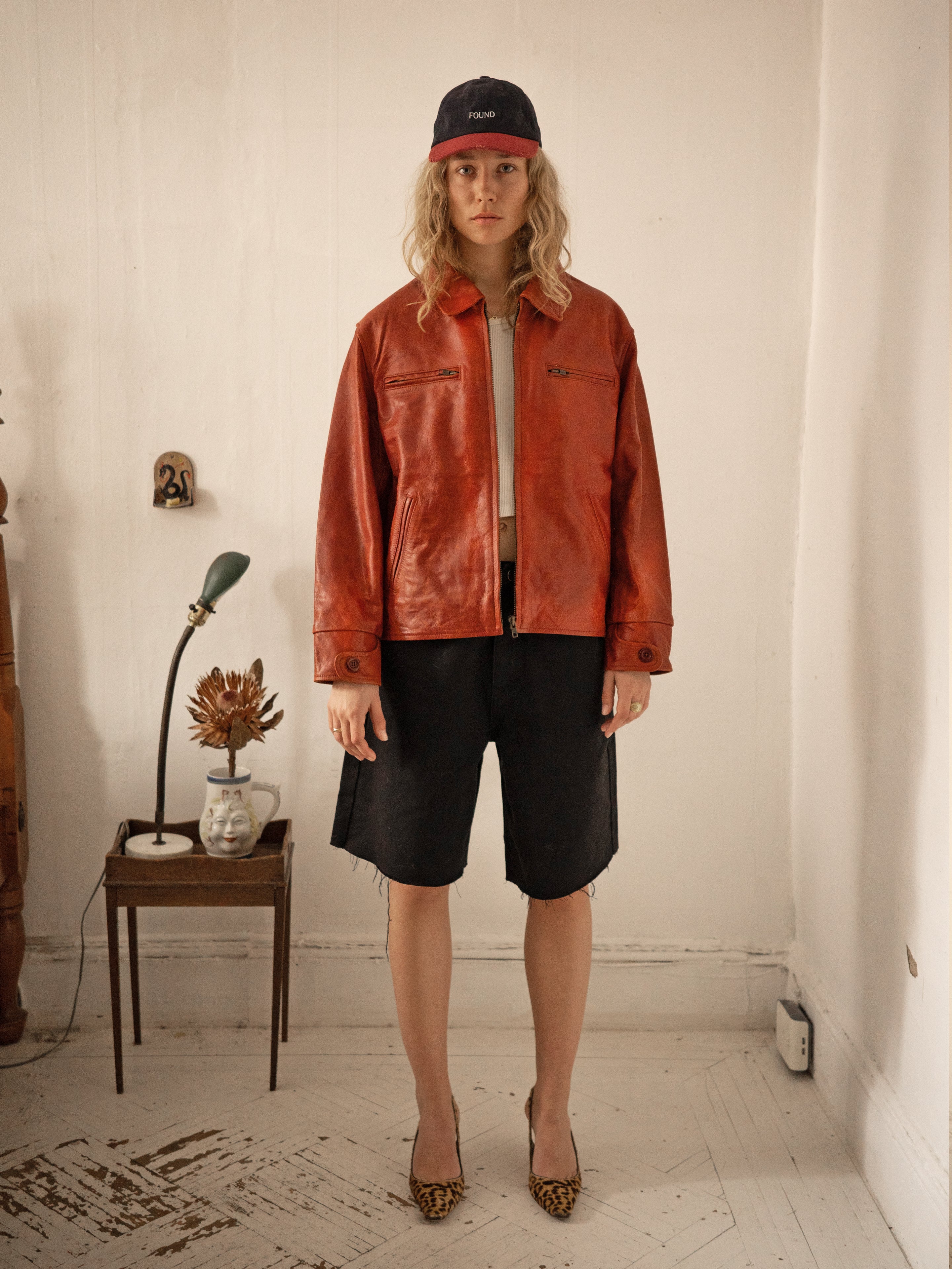 A person stands in a room wearing a FOUND Marlboro Sun Faded Leather Racer Jacket and black cap, paired with black shorts and leopard print shoes. A small table on the left holds a lamp and dried flower, creating an inviting space.