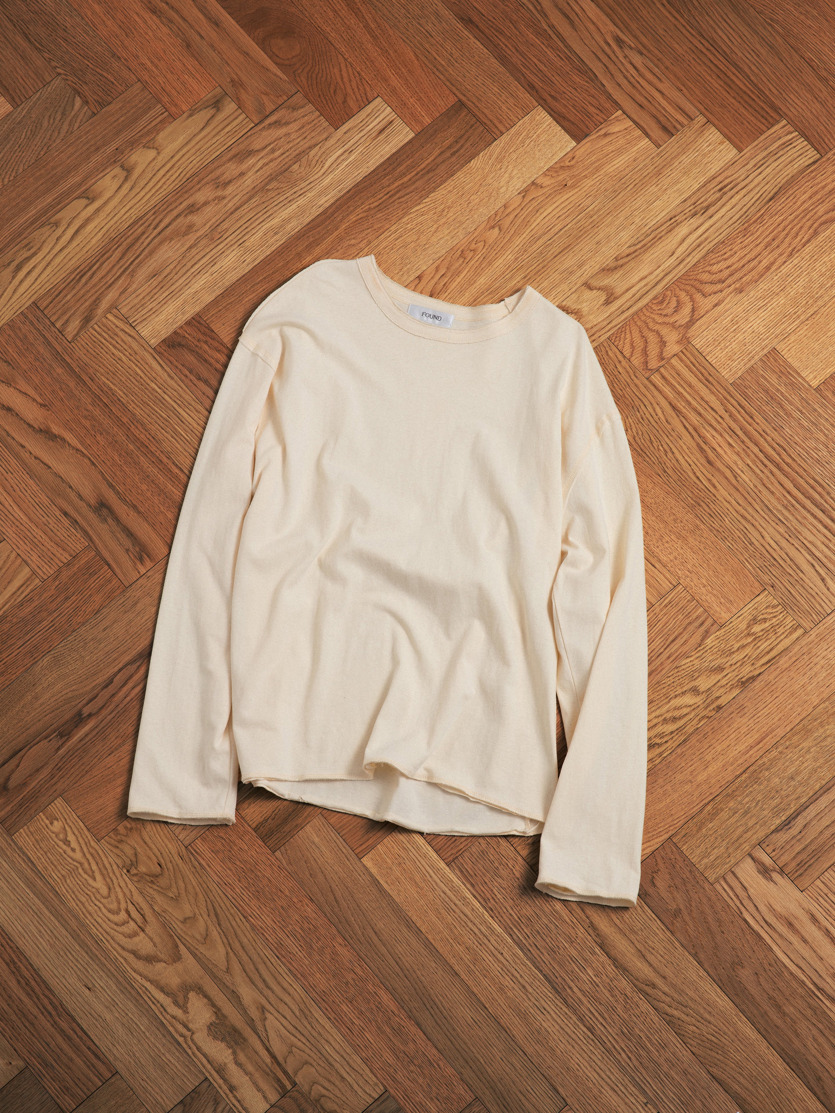 The FOUND Reversed LS Tee, a cream long sleeve with distressed neckline, is displayed flat on a wooden herringbone-patterned floor, showcasing its casual unisex style.