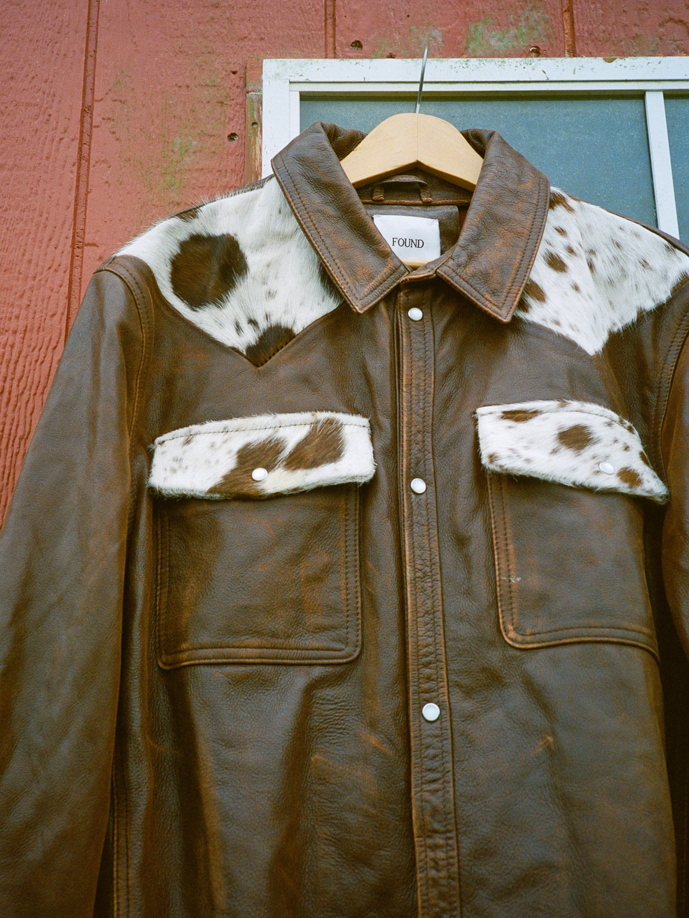 The FOUND Distressed Leather Cowhide Contrast Western Shirt, featuring cowhide-patterned accents on the collar, shoulders, and pockets, hangs on a wooden hanger before a red wooden wall and window.