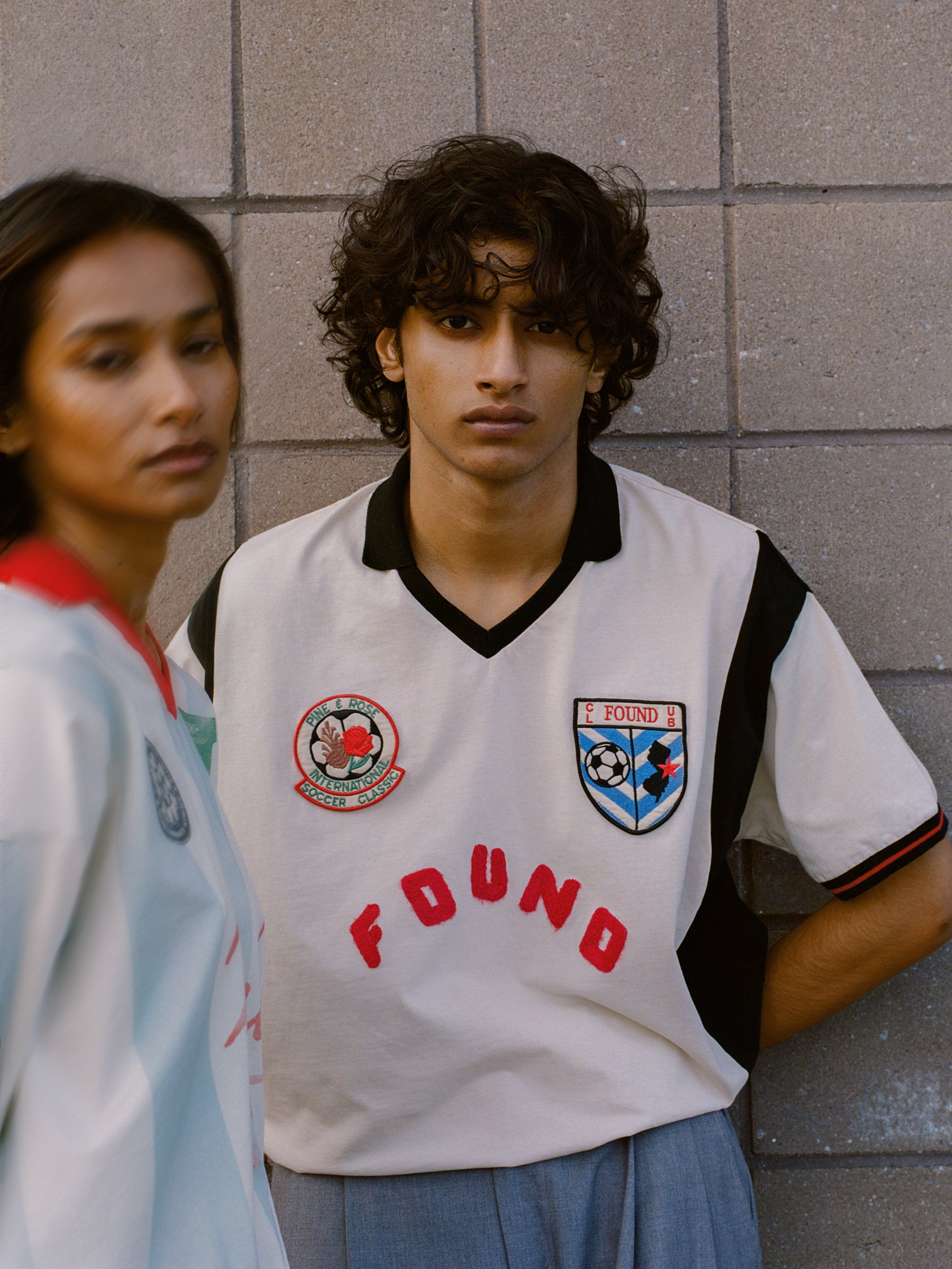 Two people stand against a tiled wall; one, sharply in focus, wears the "FOUND" Union Soccer Jersey reminiscent of vintage football gear, while the other figure blurs into the foreground.