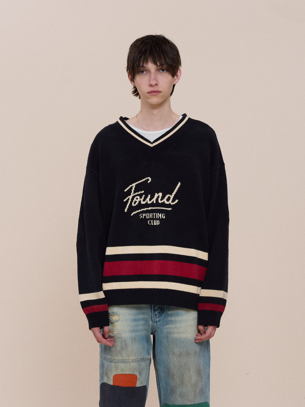 A person stands against a plain background, wearing the FOUND Sport Club Varsity Sweater with red and white stripes over a white shirt and blue jeans with colored patches.
