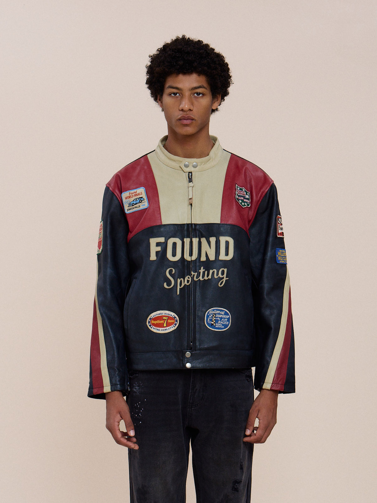 A person stands facing forward in the Sport Racer Jacket by FOUND, featuring patches and black jeans, set against a plain light background.