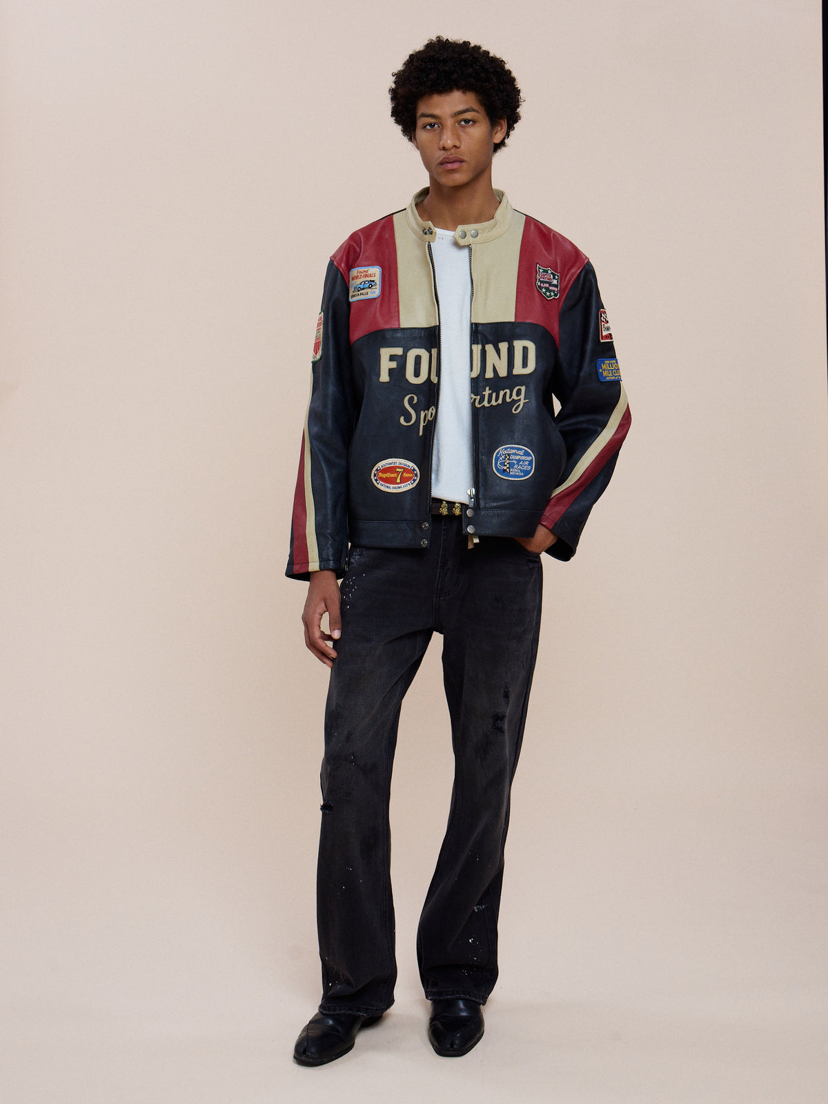 A person stands against a plain background wearing the Sport Racer Jacket by FOUND, along with a white shirt, black jeans, and black shoes.