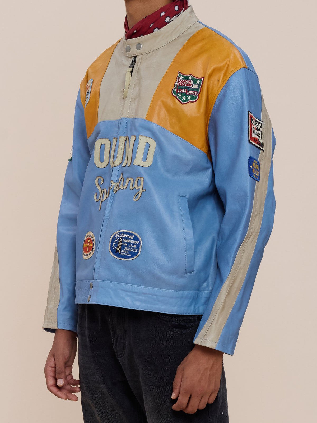 A person wears a blue and yellow Sport Racer Jacket by FOUND with racing patches, over a red shirt with white polka dots.