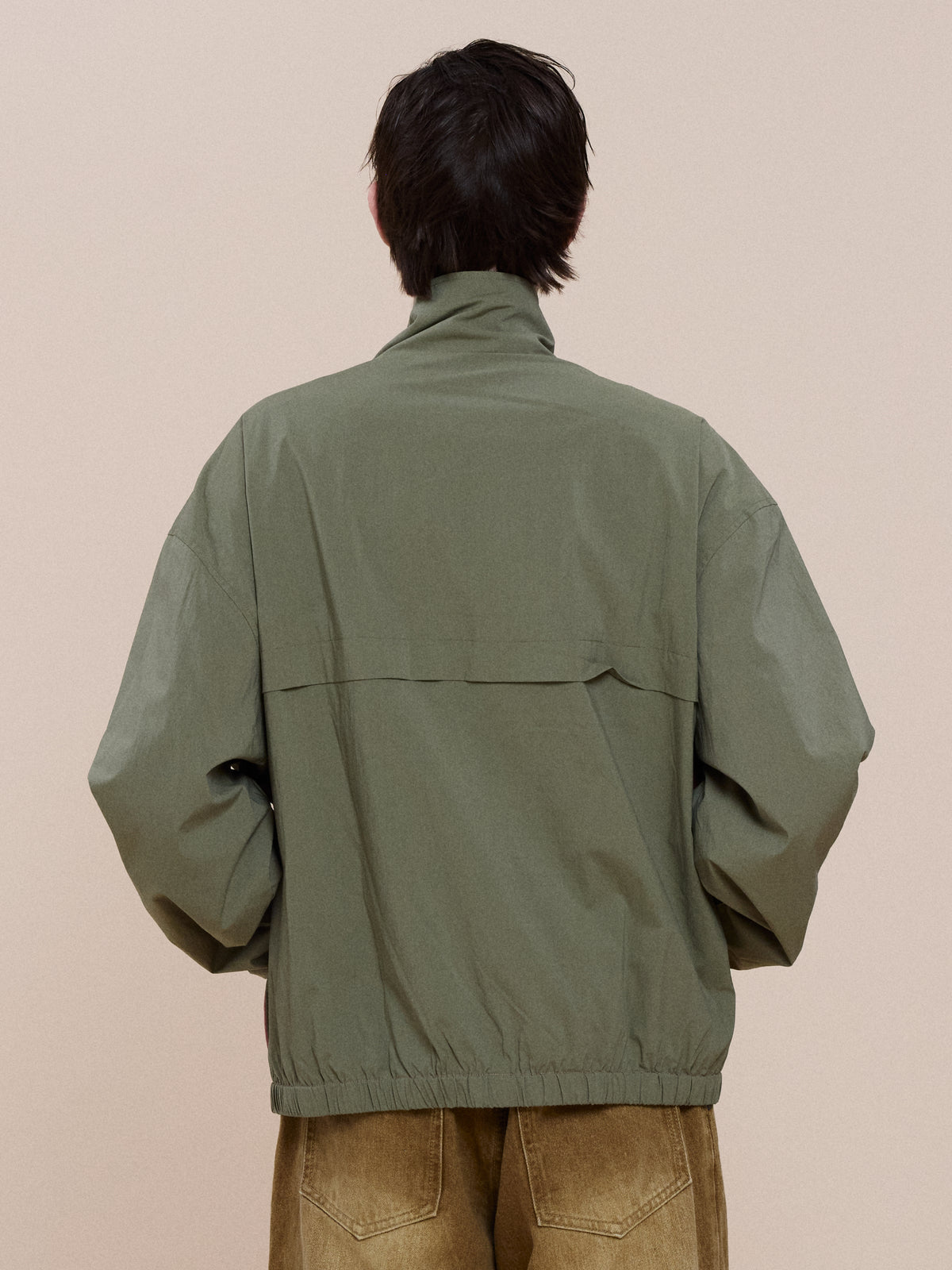 Person wearing the FOUND Sport Windbreaker Jacket in olive green, shown from the back with elastic cuffs and hem, against a plain beige background.
