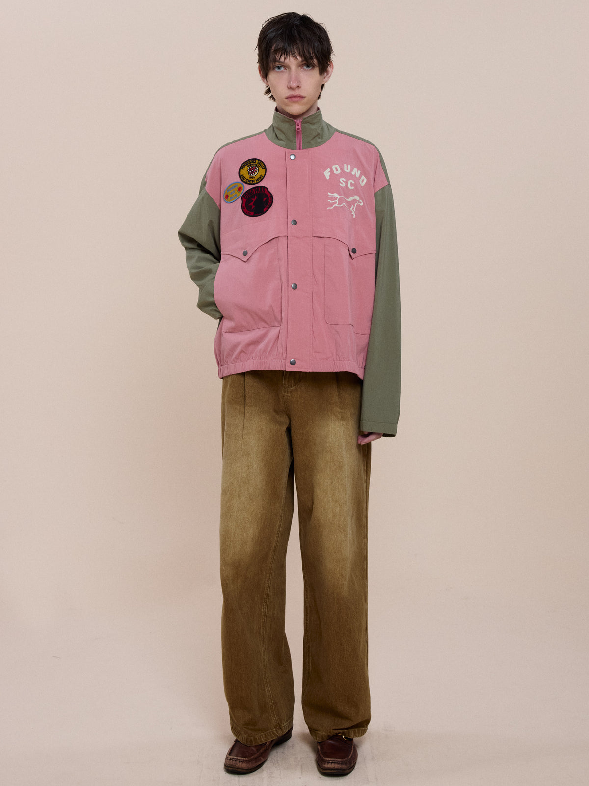 Person stands against a plain background, wearing the FOUND Sport Windbreaker Jacket in green and pink with patches, wide tan pants, and brown shoes.