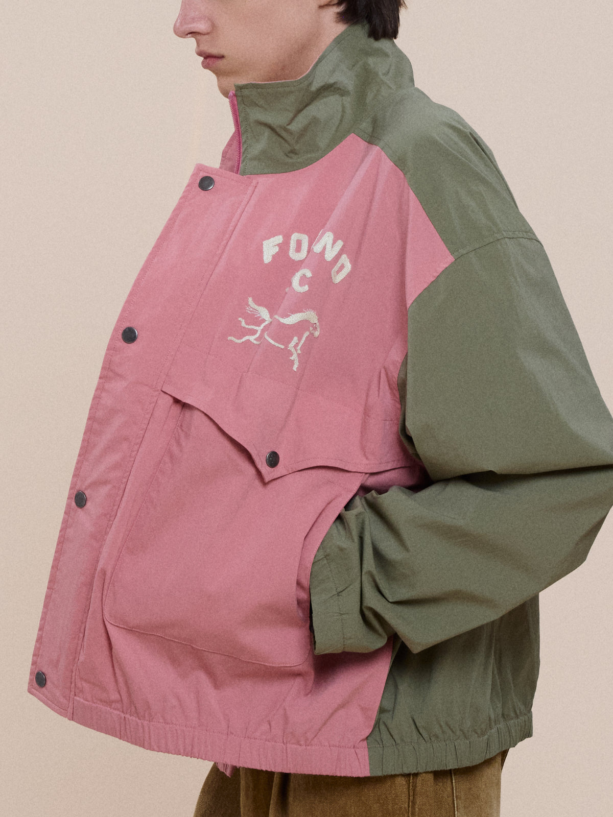 Person wearing a green and pink Sport Windbreaker Jacket by FOUND with “FOND C” and horse graphic on chest, hand in pocket, facing left.