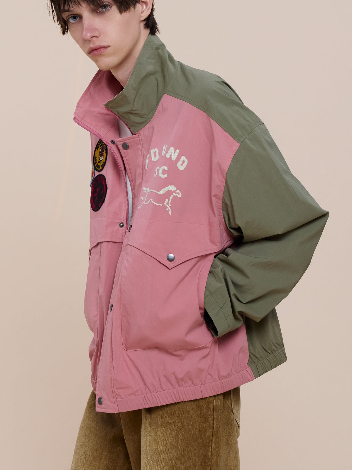 Model wears the FOUND Sport Windbreaker Jacket in pink and olive green, featuring patches and embroidery, hands in pockets against a beige background.