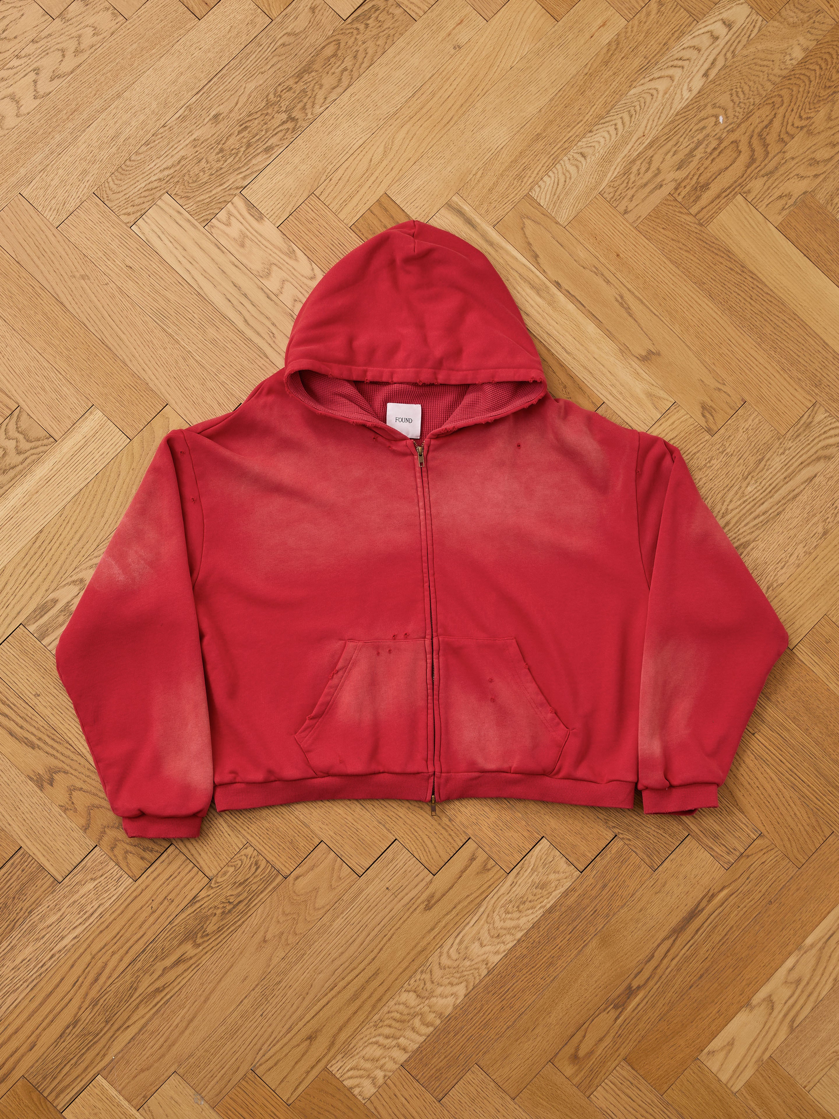 The FOUND Distressed Thermal Lined Zip Up Hoodie features a unisex relaxed fit, red color with faded, discolored areas, front pockets, and a hood. It's shown laid flat on a wooden parquet floor.