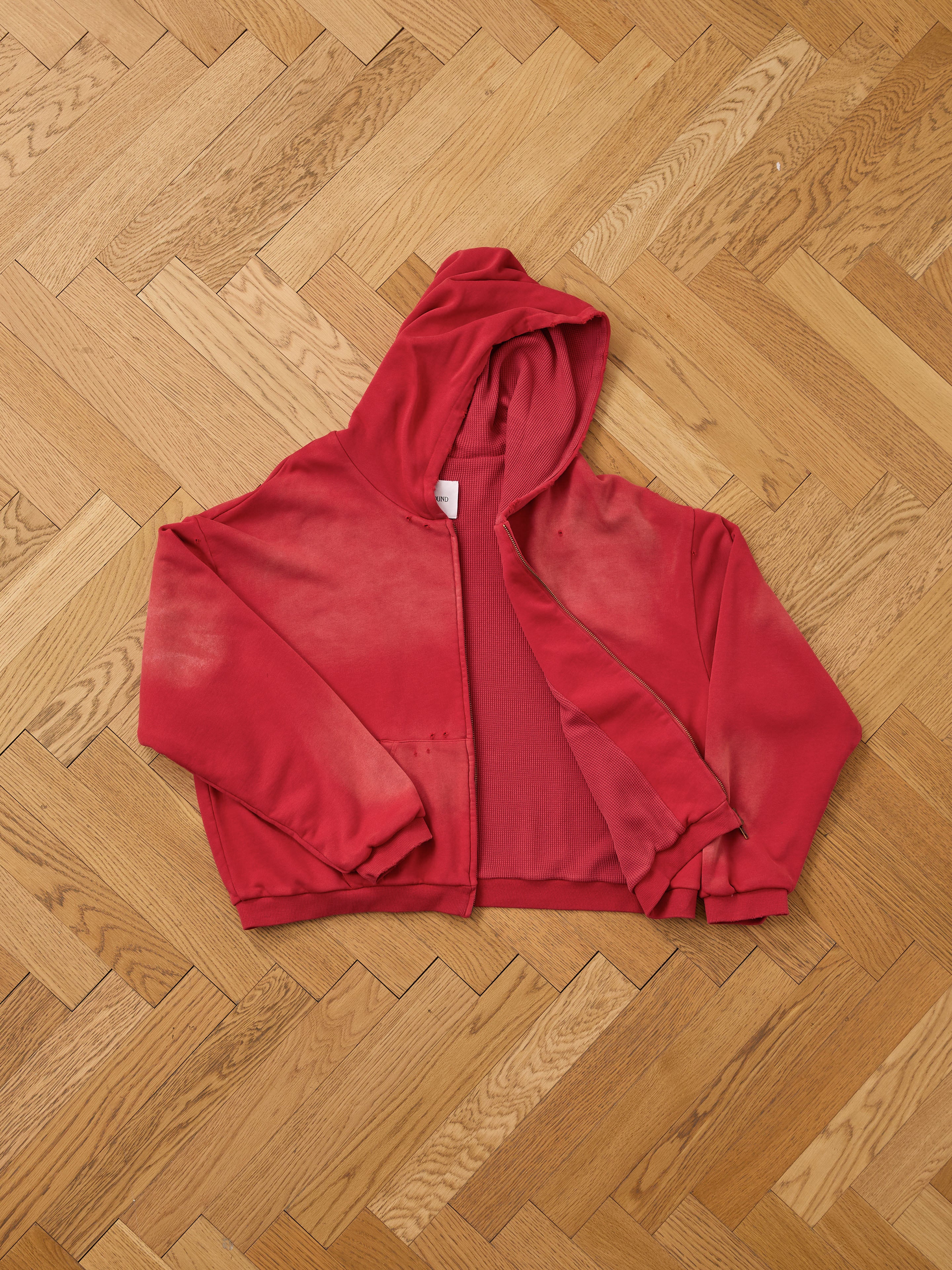 The FOUND Distressed Thermal Lined Zip Up Hoodie in red features a relaxed fit with faded and discolored details, displayed on a light wooden herringbone floor.